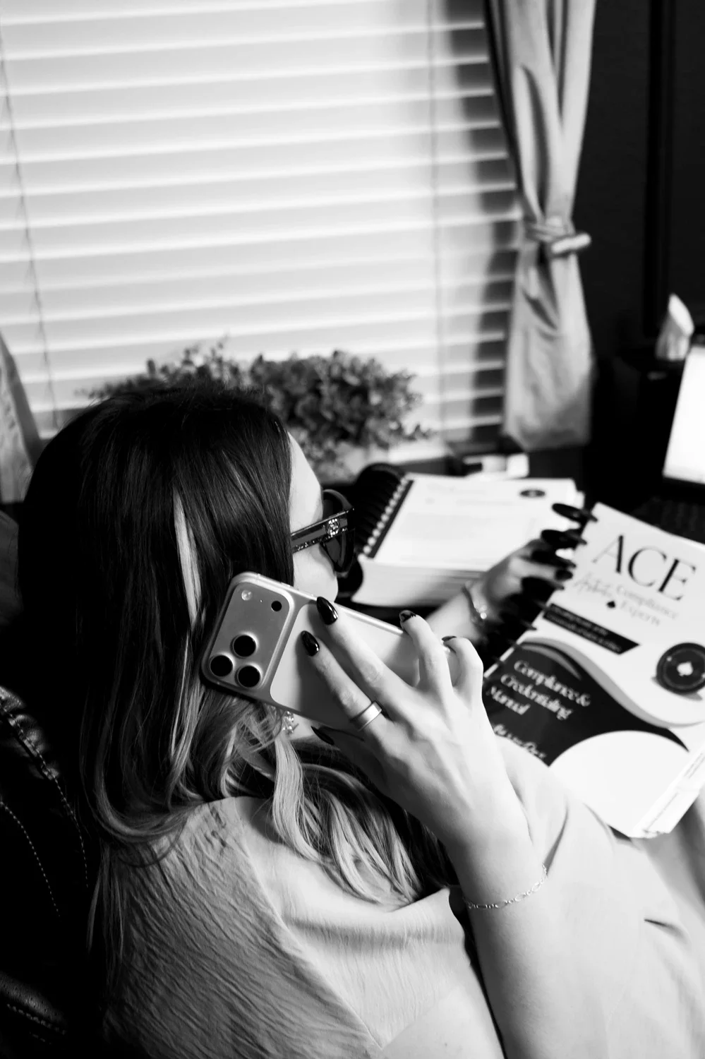 Black-and-white image of a focused woman on the phone at a desk, reviewing documents beside a laptop.