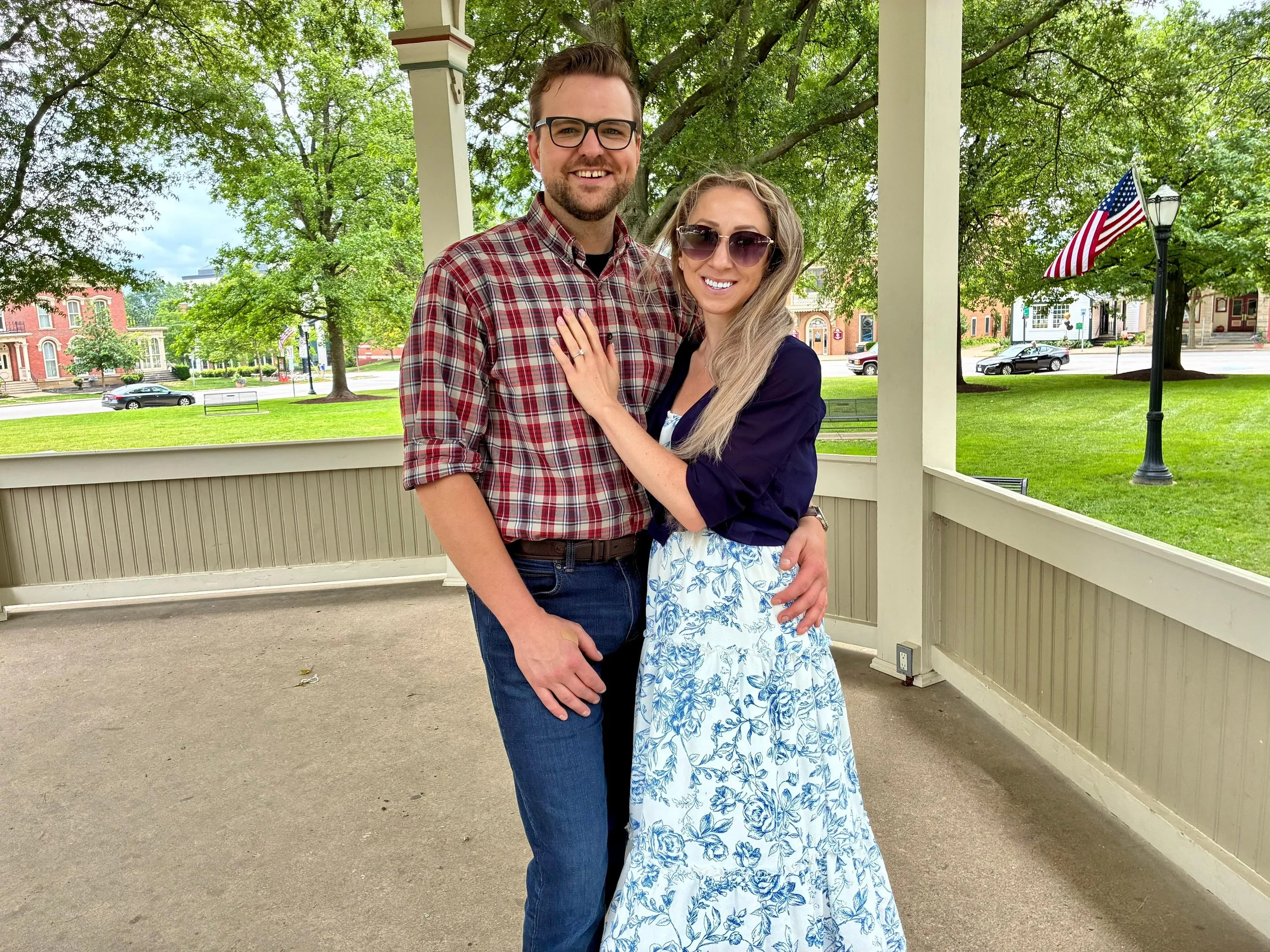 A smiling young couple standing on a porch with green trees behind them, an American flag on a pole, and residential houses visible in the background.