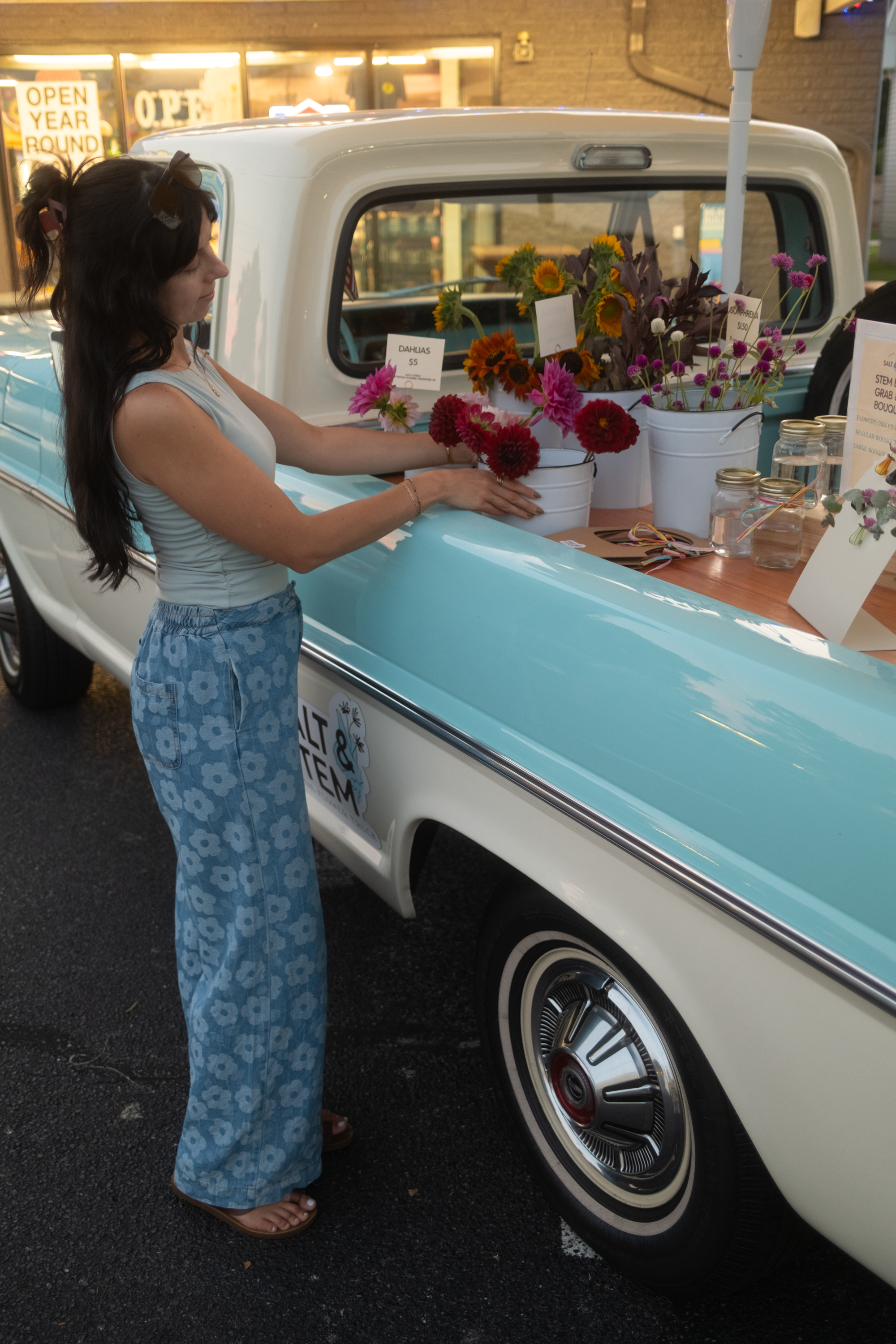 A woman with long dark hair, wearing a sleeveless white top and blue floral pants, is holding a pink flower. She is standing next to a vintage white pickup truck with turquoise accents, decorated with buckets of colorful sunflowers and other flowers. There are signs indicating flower prices and a small display of jars and paper, suggesting a flower stand or sale.