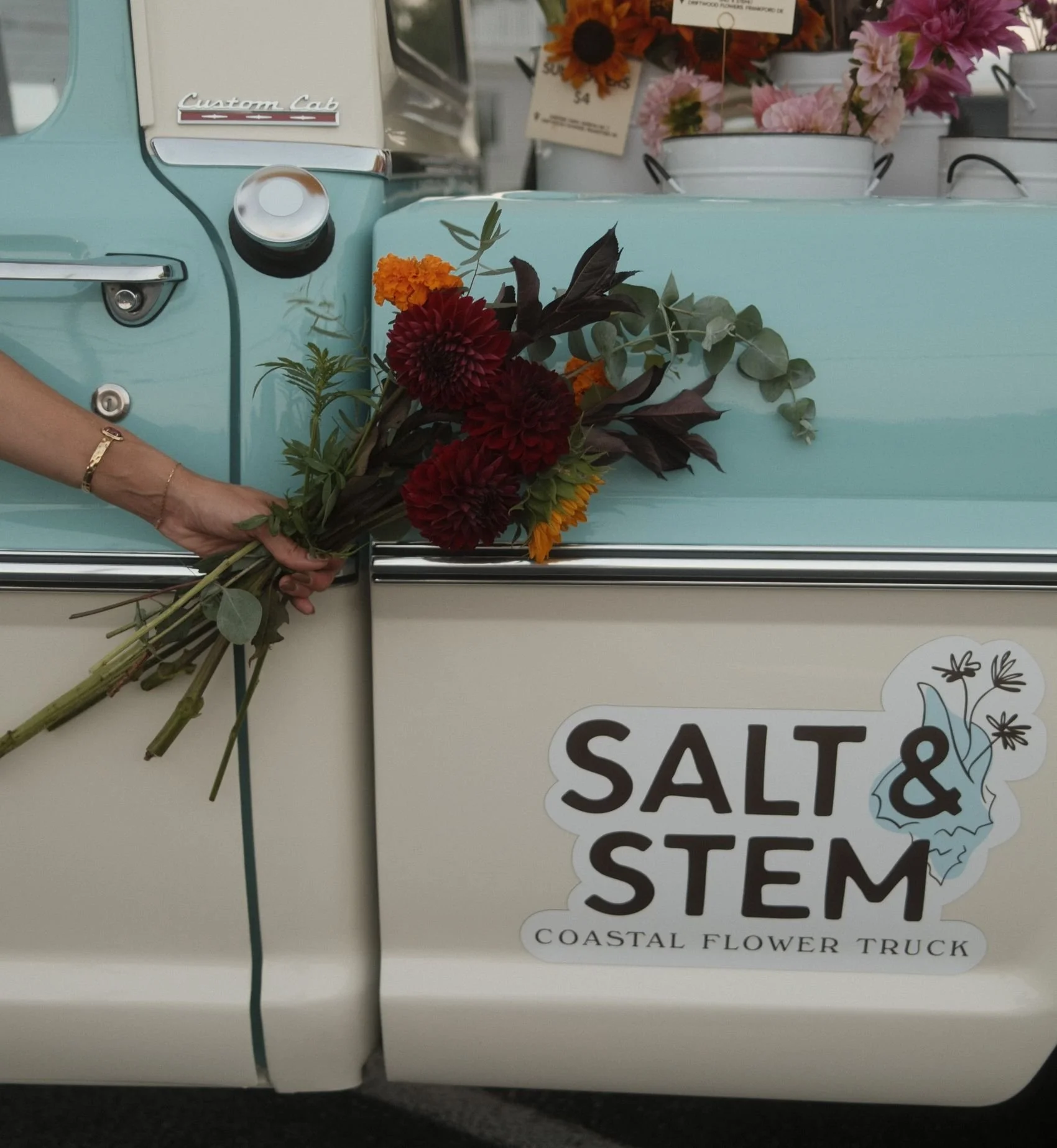 Close-up of a pastel blue coastal flower truck with a bouquet of red, orange, and purple flowers, and white buckets of flowers in the background. Sticker on the truck reads "Salt & Stem Coastal Flower Truck."