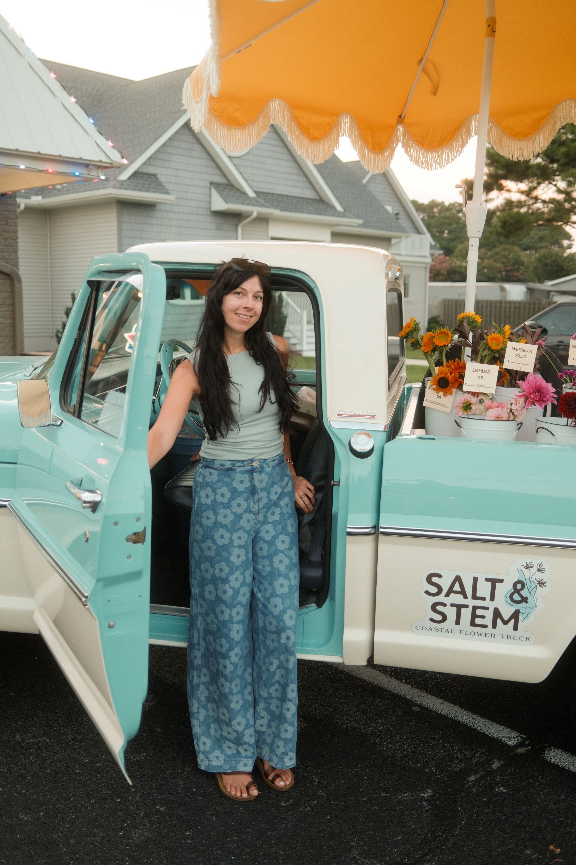 A woman standing in the doorway of a vintage teal and white truck labeled Salt & Stem Coastal Flower Truck, with a amber-yellow umbrella overhead and a basket of sunflowers and pink flowers with price tags nearby. Residential houses are seen in the background during sunset.