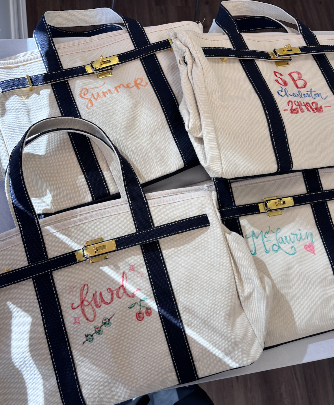 Four white canvas tote bags with black handles and gold clasps, each personalized with colorful handwritten names and designs, placed on a table.