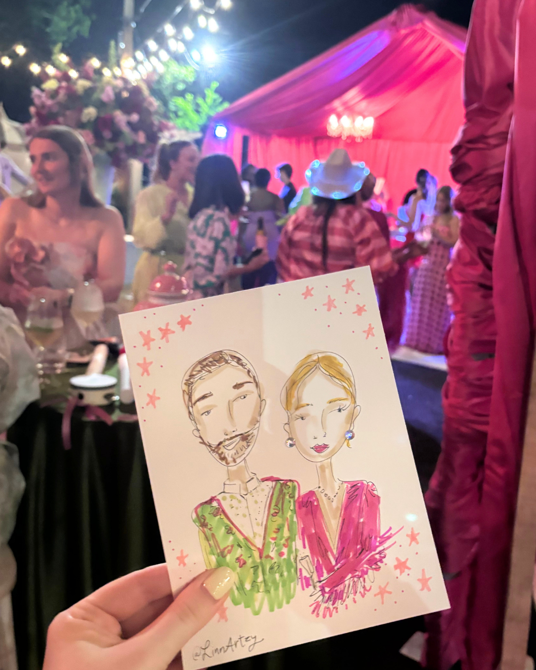 A person holding a colorful caricature of a man with a beard and woman with earrings at a festive event with string lights and a pink tent in the background.