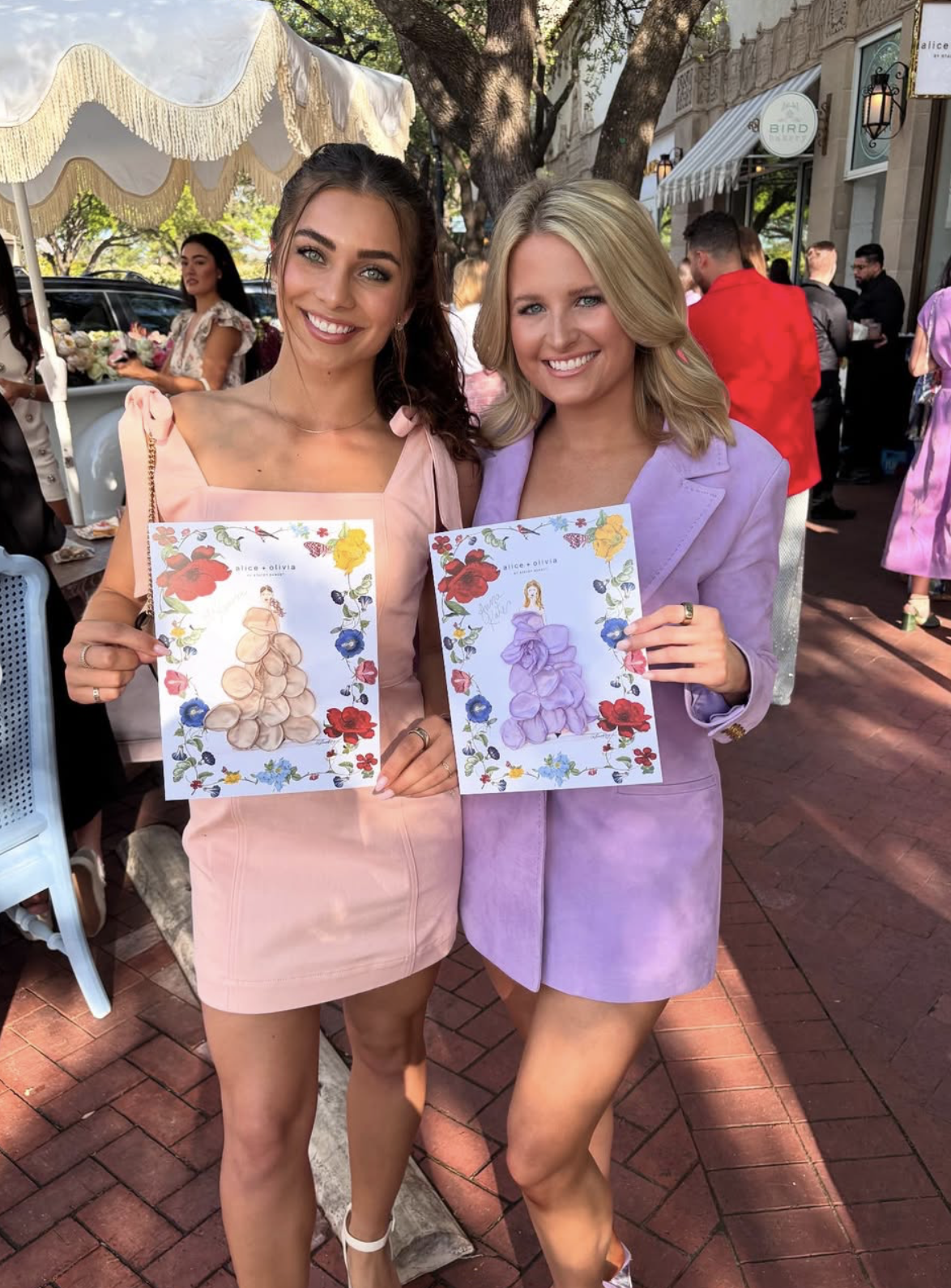 Two women wearing pastel-colored dresses are standing outdoors on a brick sidewalk, each holding illustrated artwork with floral borders and fashion sketches. They are smiling and appear to be at an event with other people and trees in the background.