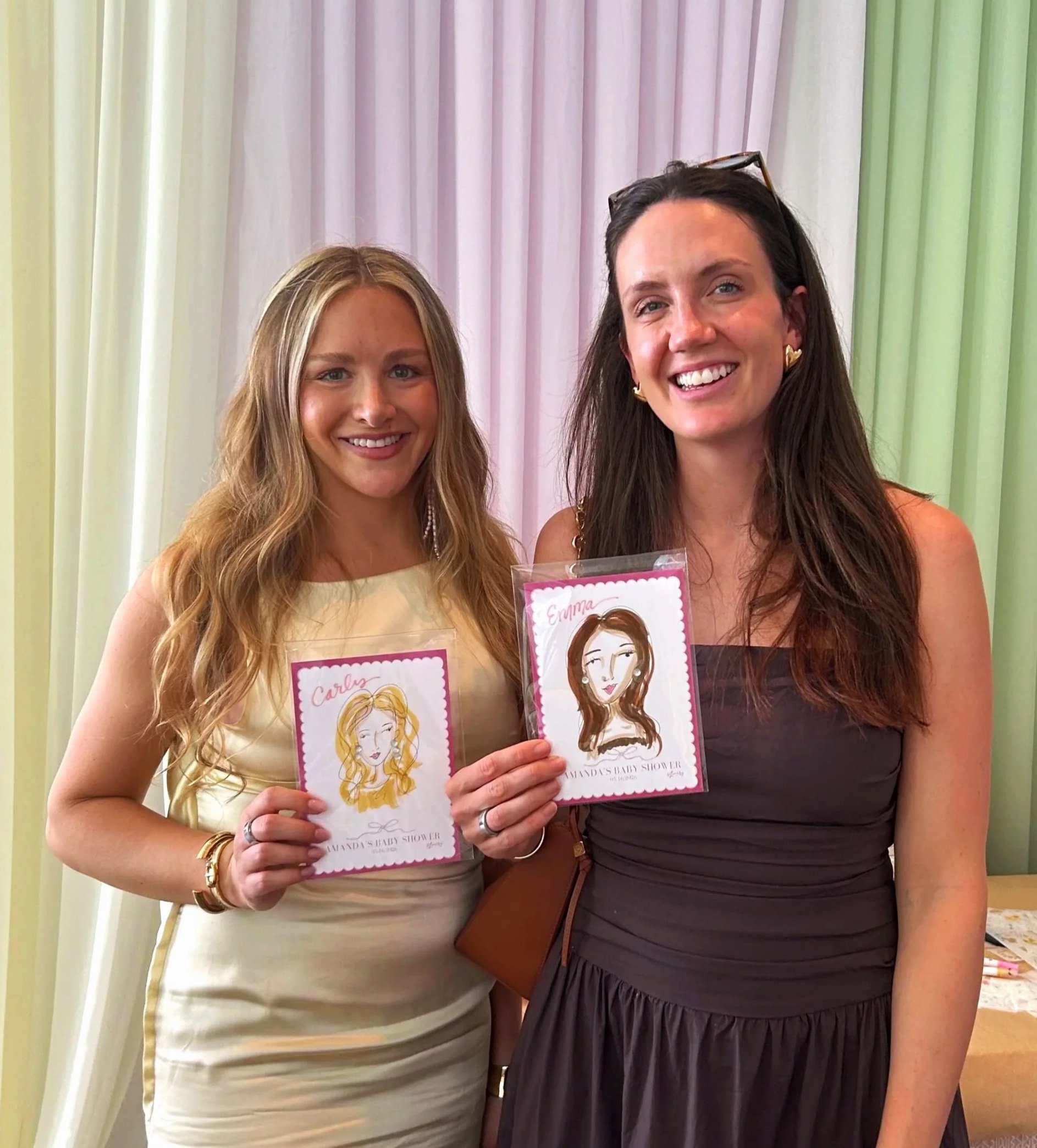 Two women smiling and holding illustrated cards, standing in front of a curtain with pastel colors.