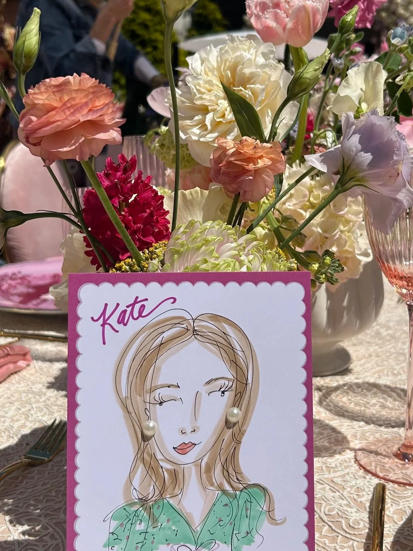 A floral centerpiece with pink, peach, white, and cream-colored flowers in a glass vase. In the foreground, there is a drawing of a woman's face with pearl earrings, blonde hair, and a green top, on a pink card that appears to be a party or celebrati
