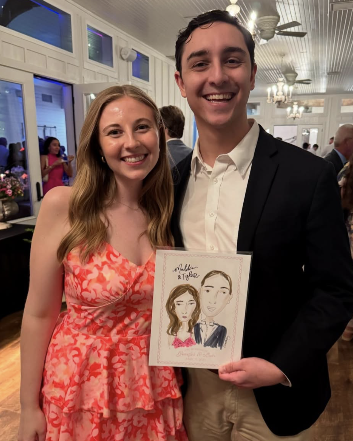 A smiling woman with long blonde hair in a pink floral dress standing next to a smiling man in a black suit jacket and white shirt, holding a caricature drawing of themselves at a social event, with other guests and chandeliers visible in the backgro