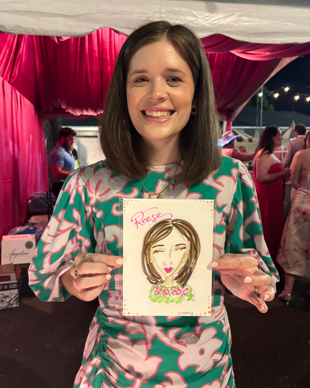 A woman smiling and holding a colorful illustration of herself with the name 'Reese' written on it. She is at an outdoor event with pink and red drapery, surrounded by other people in evening attire.