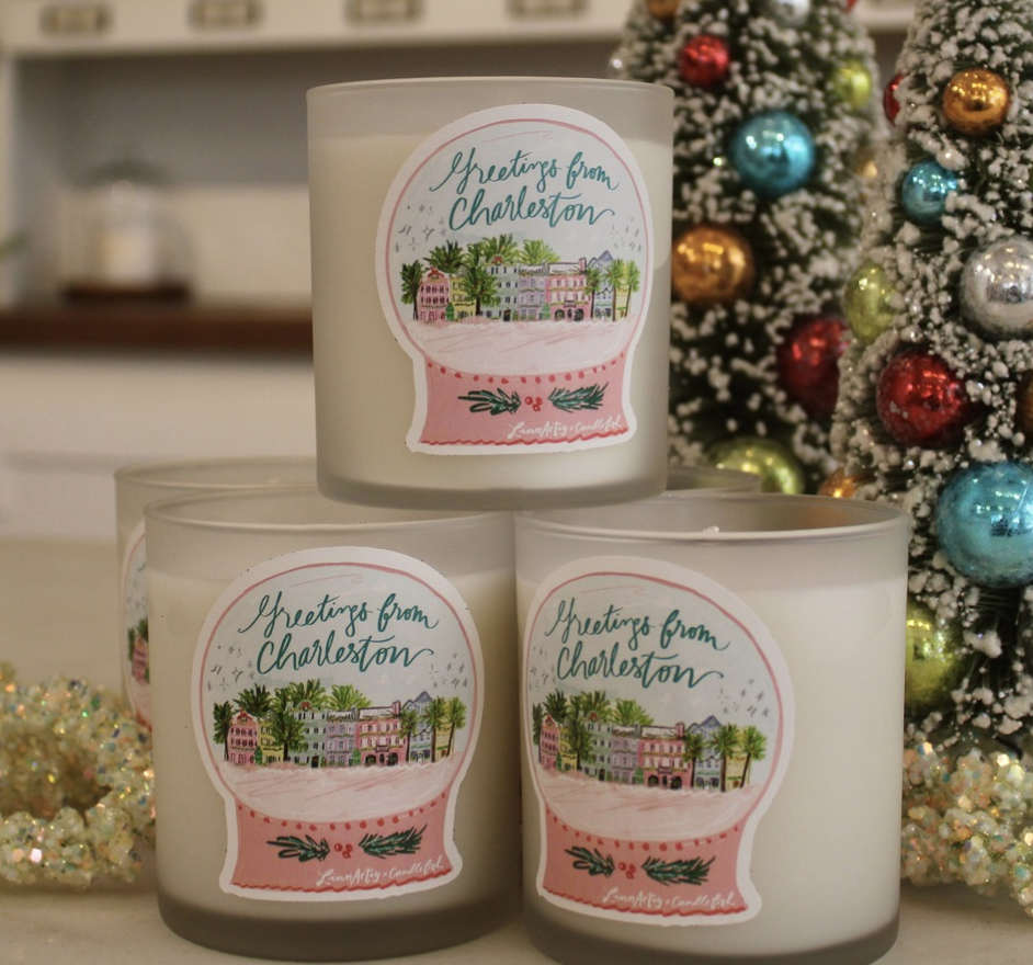 Three white candles with 'Greetings from Charleston' stickers featuring a colorful illustration of Charleston, South Carolina, beside a decorated Christmas tree.