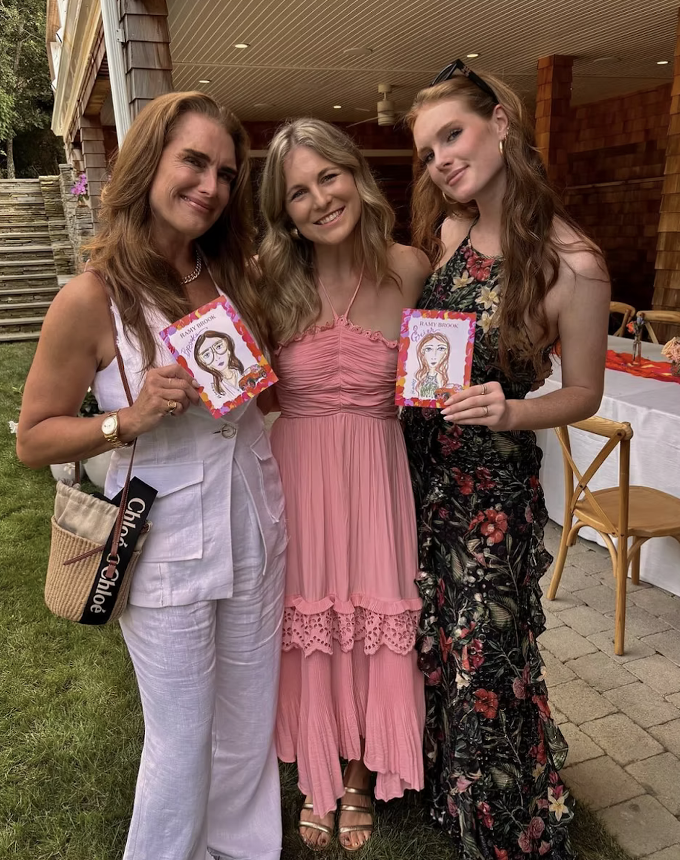 Three women standing together outdoors, smiling, two holding illustrated cards of themselves, in front of a brick building with a table and chairs nearby.