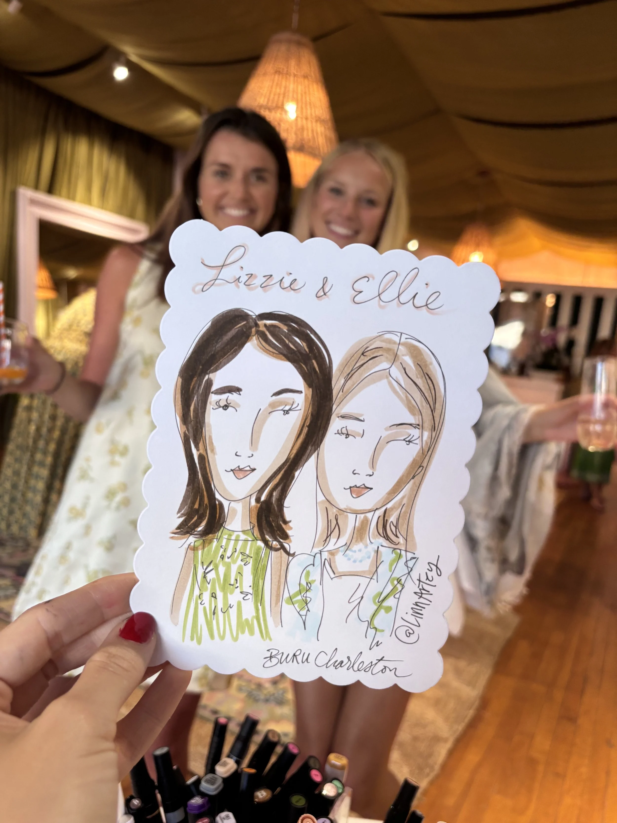 Two women smiling behind a hand-drawn caricature of their faces with the names Lizzie and Ellie written on top at an indoor event with warm lighting.