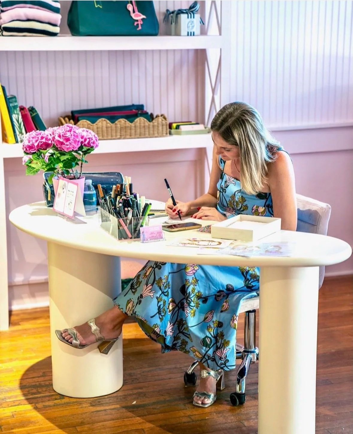 A woman sitting at a white desk with a blue floral dress, writing with a black pen, surrounded by office supplies and pink flowers in a pot, in a bright room with wooden floors and white shelves.