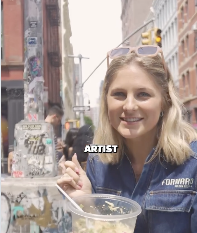 Young woman with blonde hair, sunglasses on her head, smiling, holding a spoon, sitting at a table outdoors in an urban area, with a city street and buildings in the background.