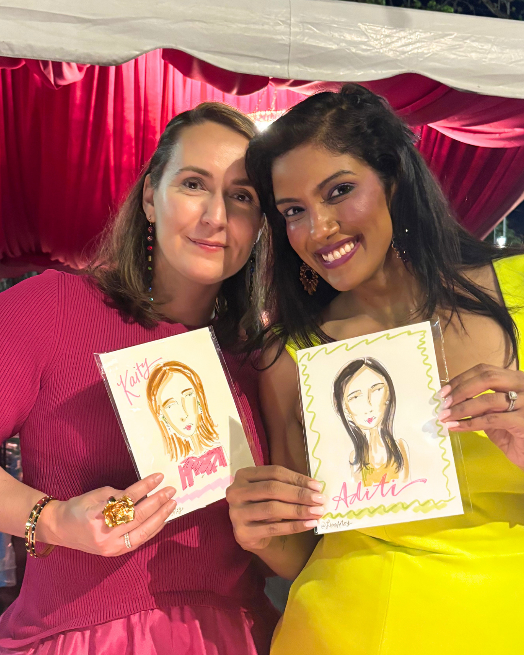 Two women smiling, holding colorful drawings of women with their names, Katie and Aditi, written on them, in front of a red curtain at an event.