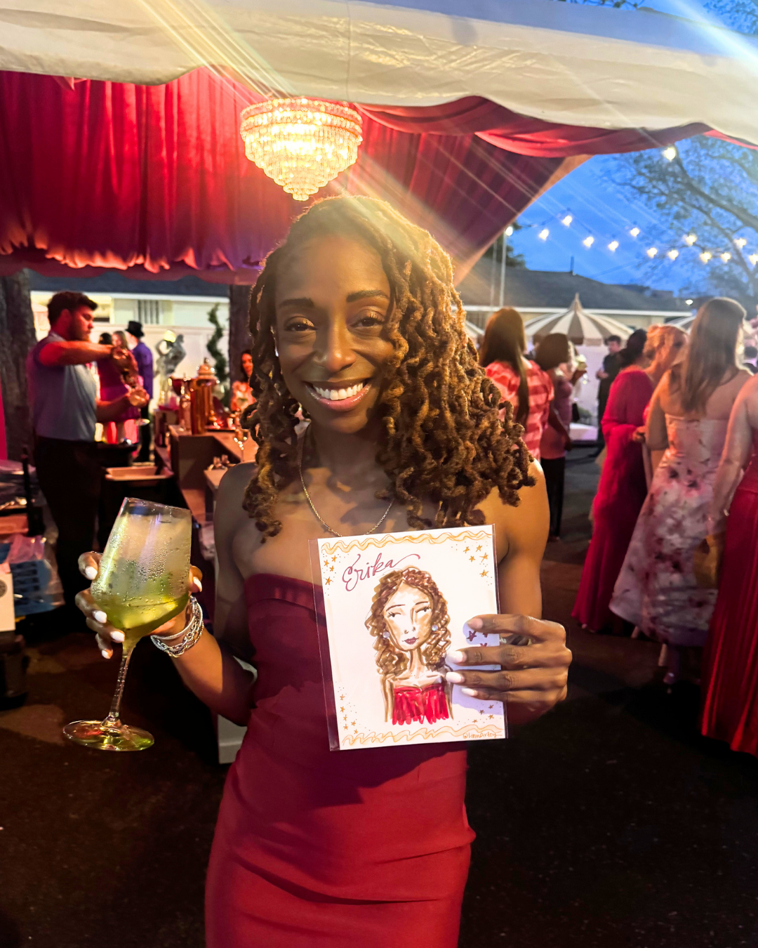 A woman with curly hair in a strapless red dress holding a glass of drink and a drawing of herself at a festive party.