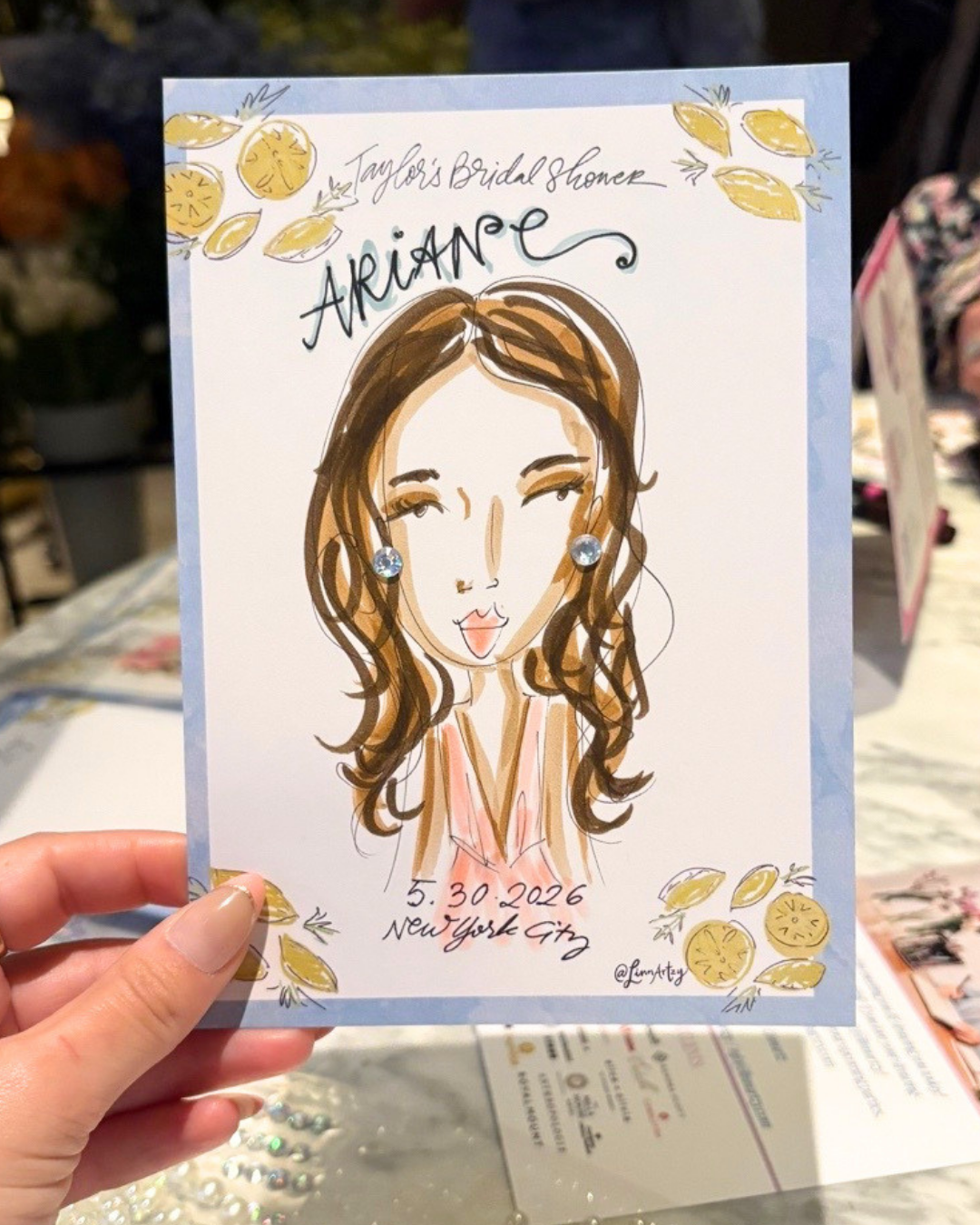 A hand holding a cartoon illustration of a woman with brown, wavy hair, wearing earrings, and sticking out her tongue, on a bridal shower card with lemon decorations. The card has the inscription "Taylor's Bridal Shower" at the top, the name "Ariane"