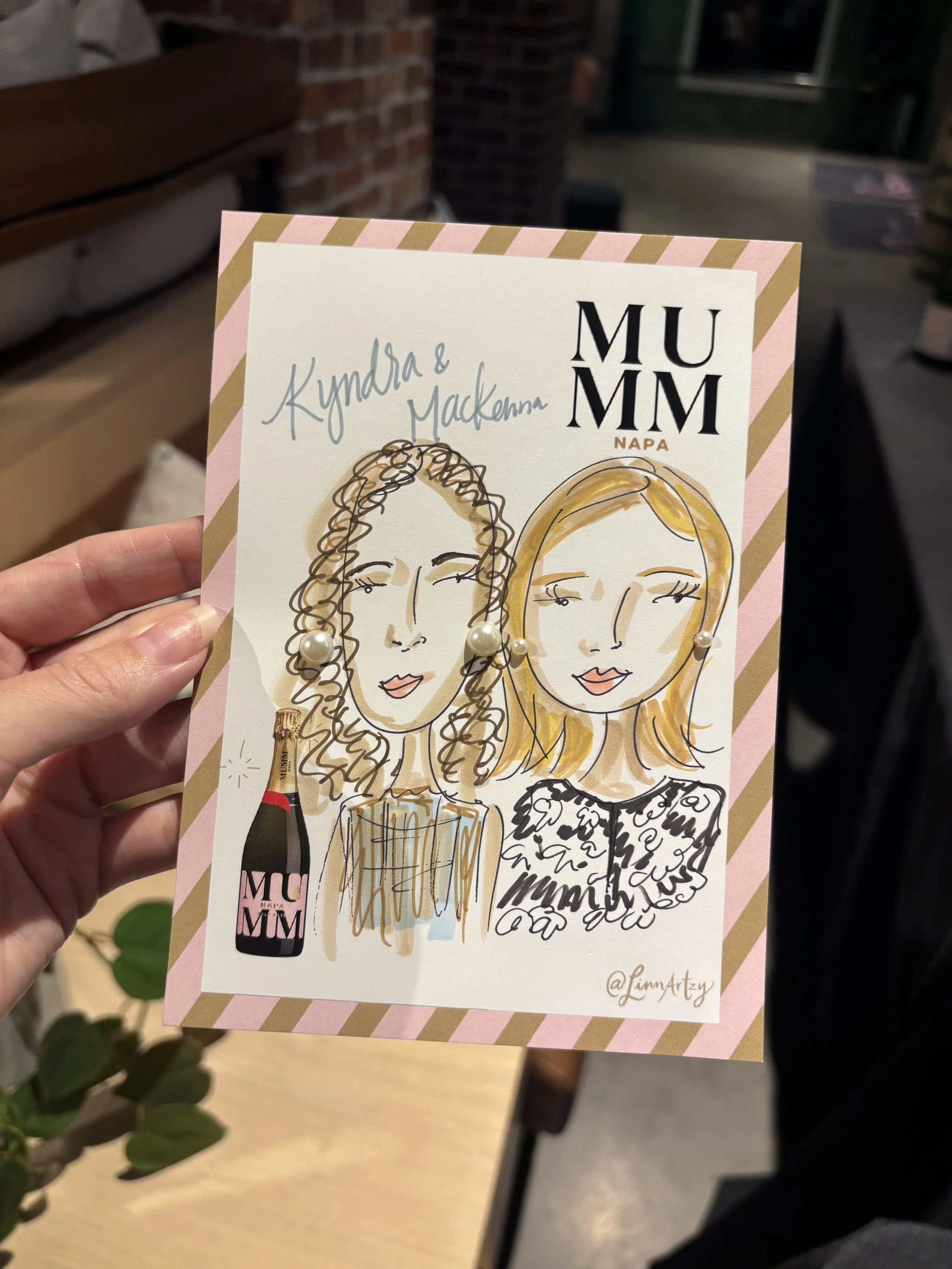 A hand holding a illustrated card featuring two women, a wine bottle, and a glass of wine. The card reads 'Kyndra & Mckenna' and 'MUMM Napa' with a signature '@LinnArtsy' at the bottom right.