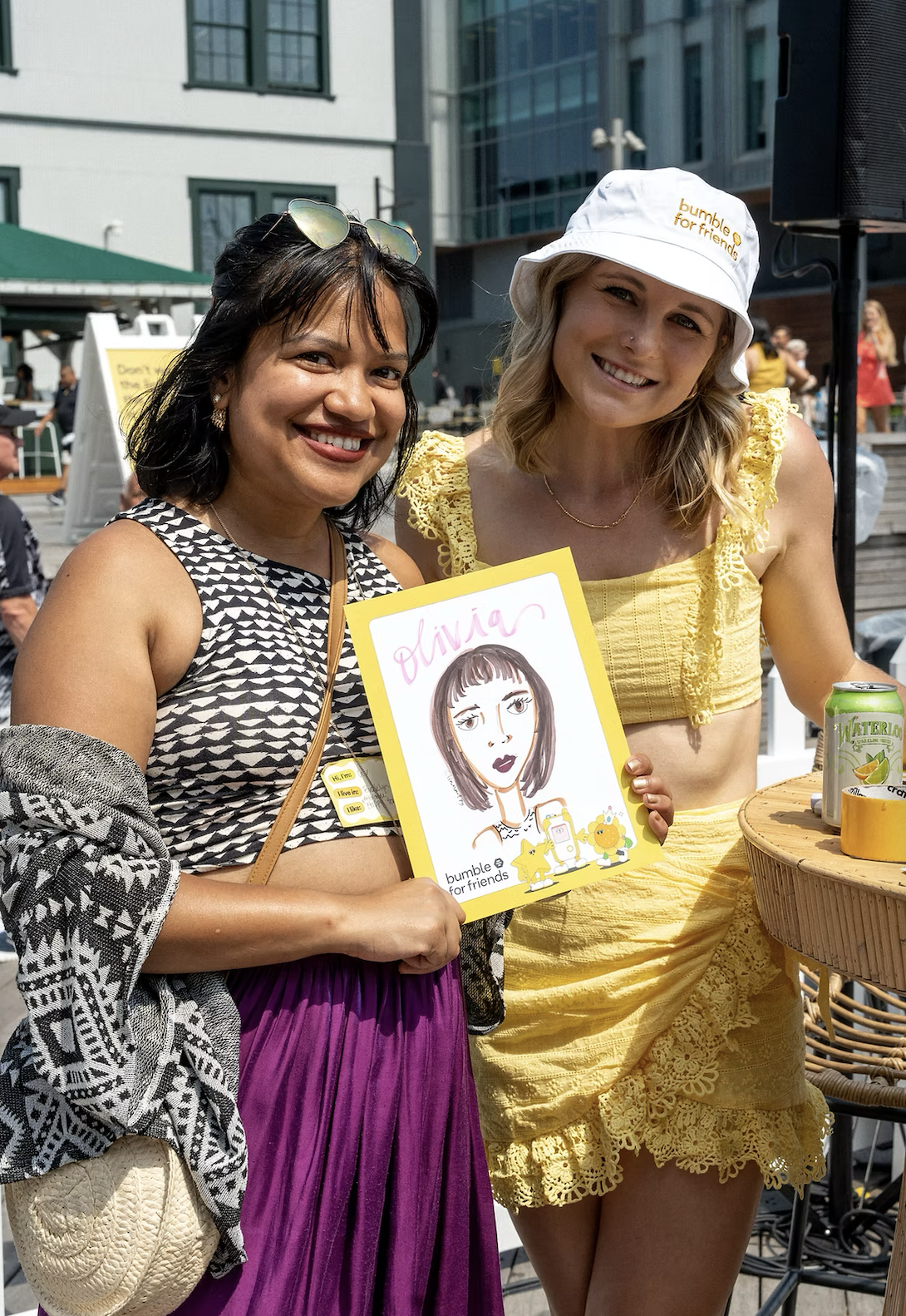 Two women smiling outdoors, one holding a colorful illustrated portrait of herself, in a sunny urban setting at a social event, with drinks and other people in the background.