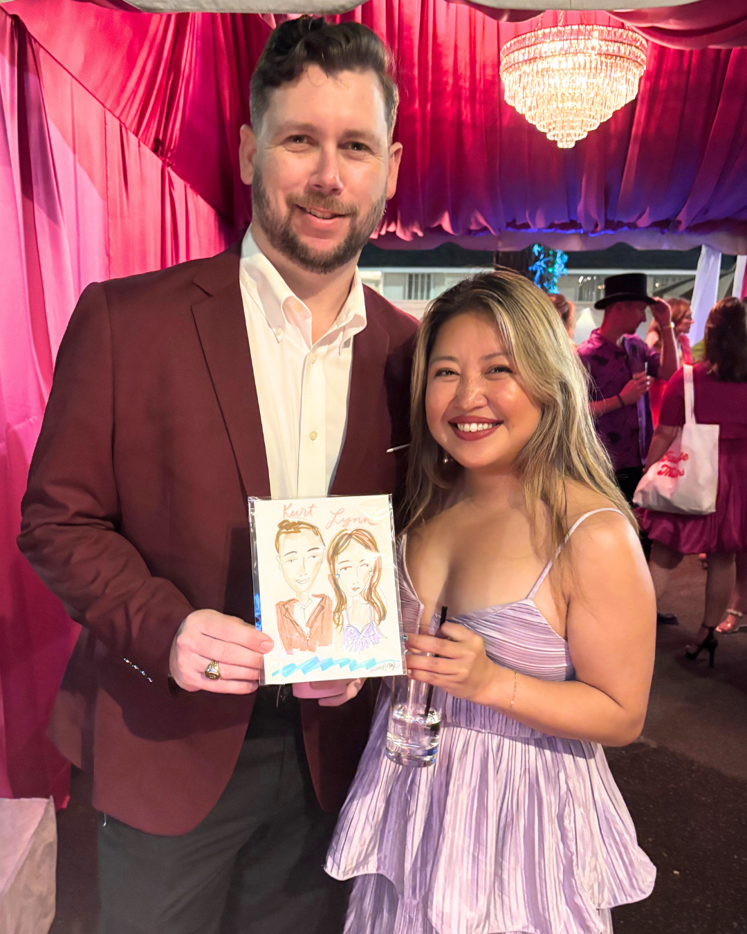 A man and woman smiling and posing together at a party, holding a drawing of themselves. The man has short dark hair, a beard, and is wearing a maroon blazer with a white shirt. The woman has shoulder-length light brown hair with blonde highlights an
