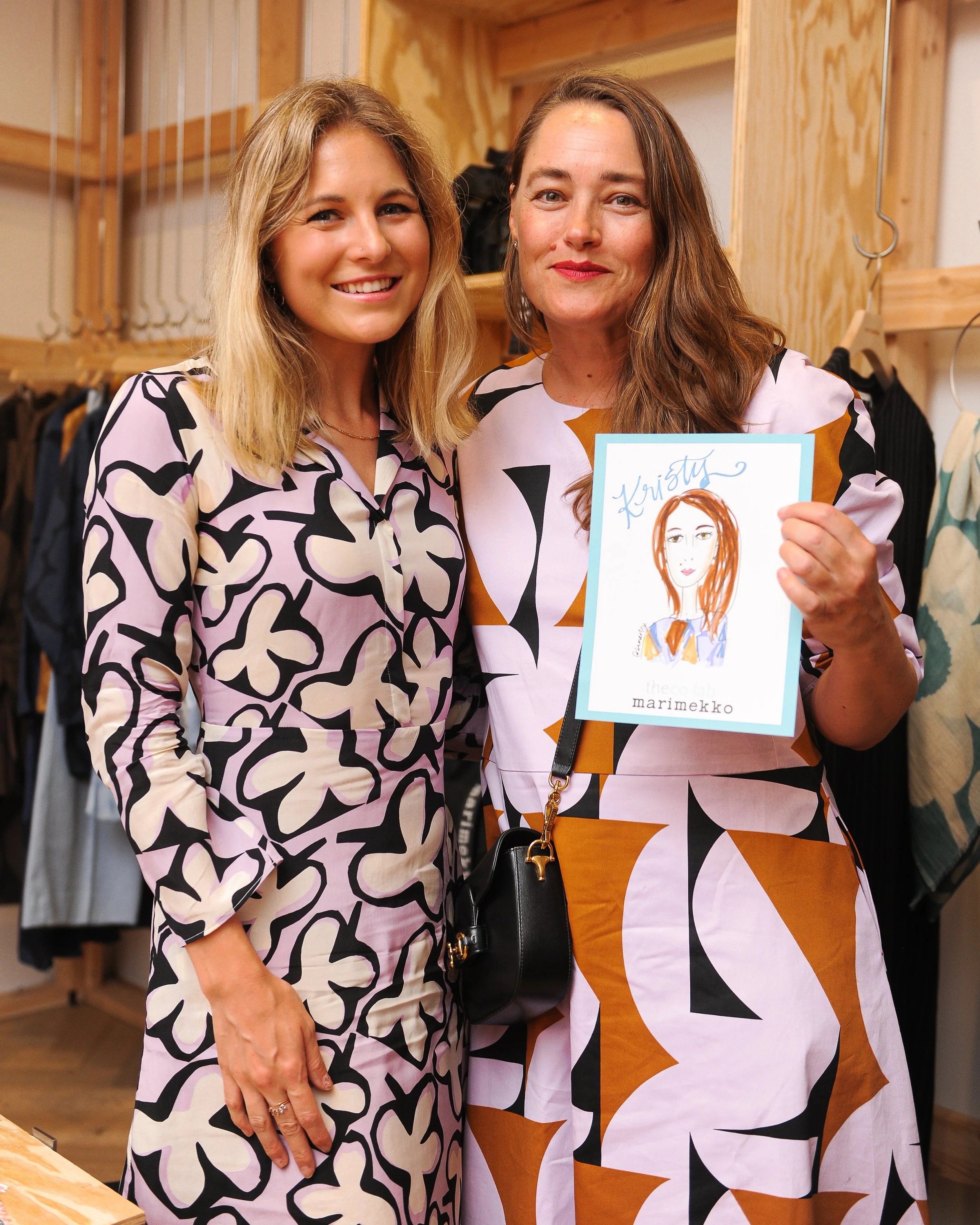 Two women smiling and standing close together inside a clothing store. One woman holds a framed illustration of a woman with red hair, labeled 'Kristy' and 'marimekko'. Both women are wearing patterned dresses.