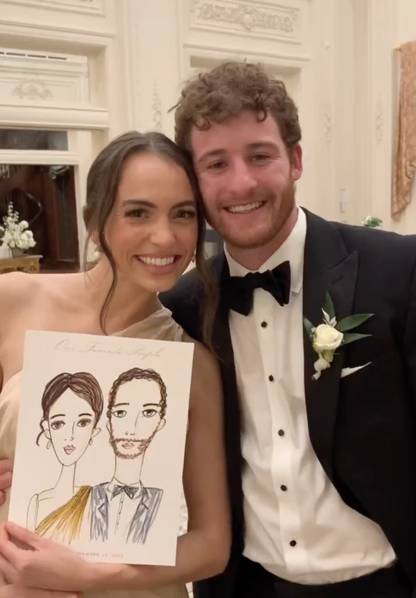 A smiling woman in an elegant dress holding a drawing of herself and a man, next to a smiling man in a tuxedo with a boutonniere, in a decorated room with ornate white walls and a mirror.