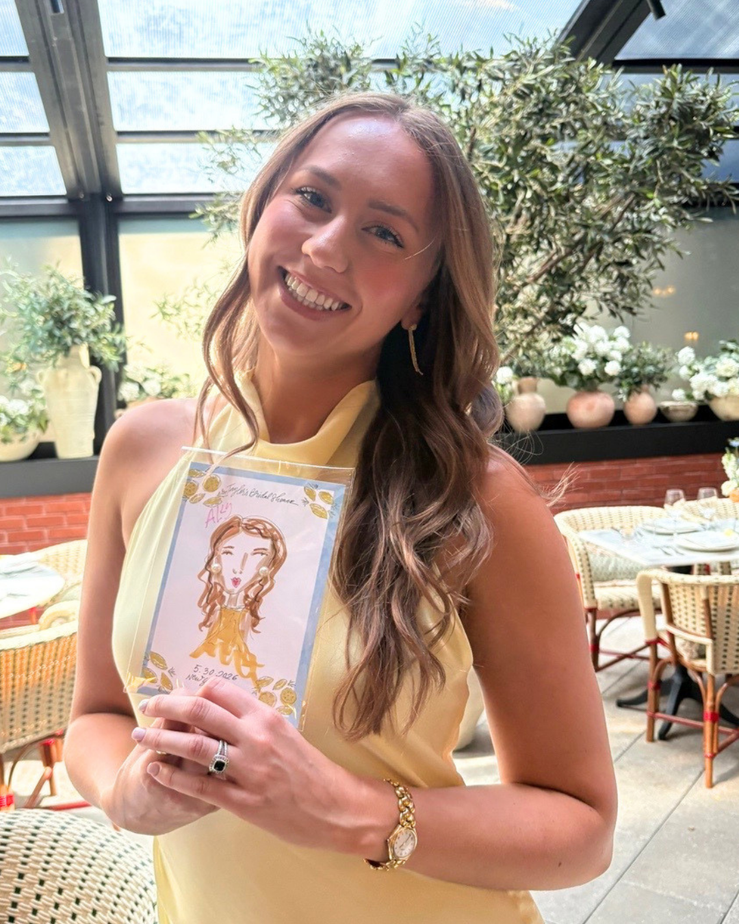 A woman with long wavy brown hair smiling and holding a caricature sketch of herself inside a clear plastic sleeve. She is wearing a sleeveless yellow dress and gold jewelry, standing in a sunlit indoor or patio setting with plants and a table set fo