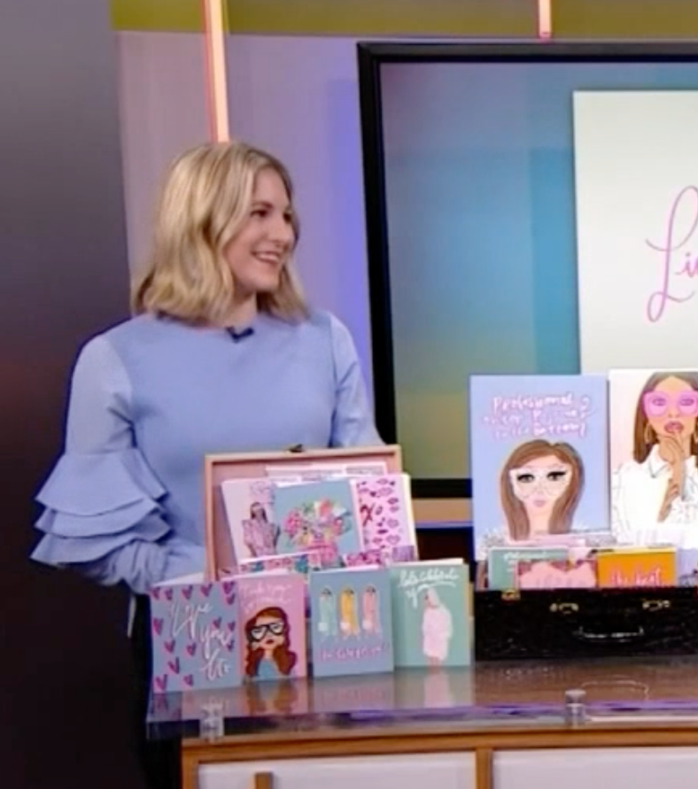 A woman standing next to a table filled with colorful illustrated books and greeting cards, in a TV studio with a large monitor displaying the word 'Lisa' in pink script.