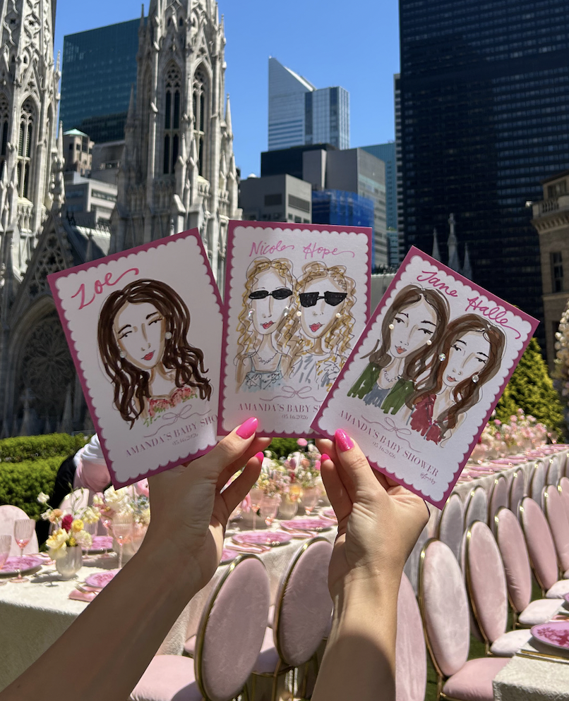 Three hand-drawn caricature cards of women with names Zoe, Nicole Hope, and Jane Hale held up in front of a cityscape with Gothic-style cathedral and skyscrapers; decorated outdoor event table with pink chairs and white flowers in the background.