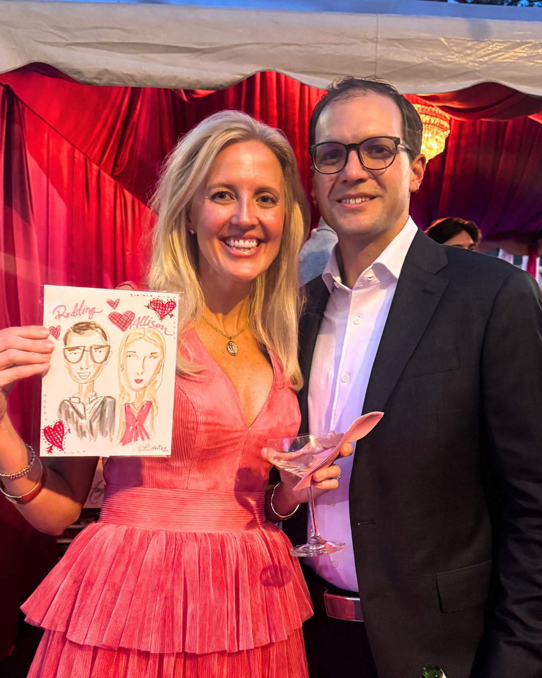 A woman and a man at a celebration, the woman holding a drawing of the pair with hearts around them and the word "Redding" and "Allison." The woman is wearing a pink dress and the man is in a black suit with glasses. They are in a venue with red curt