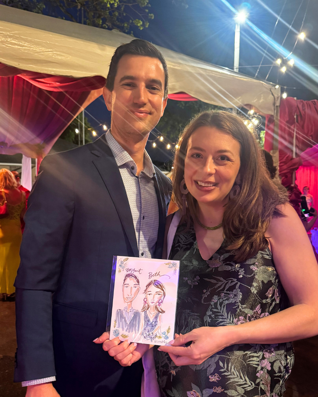 A man in a suit and a woman in a floral dress holding a drawing of themselves at an outdoor event during nighttime with string lights in the background.
