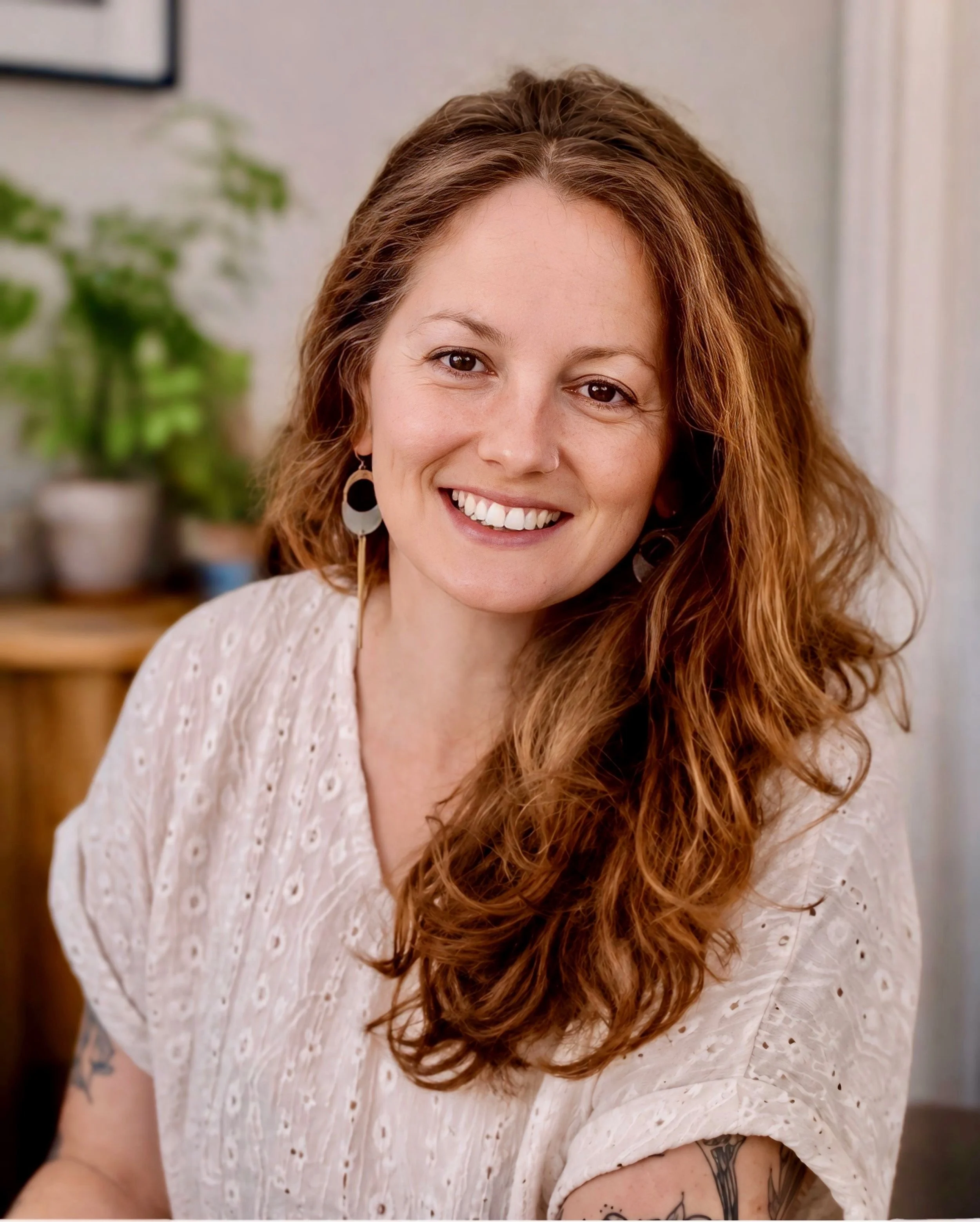 Close-up of a smiling woman with long, curly red hair wearing a light-colored, patterned top and large earrings, sitting indoors with a blurred background.