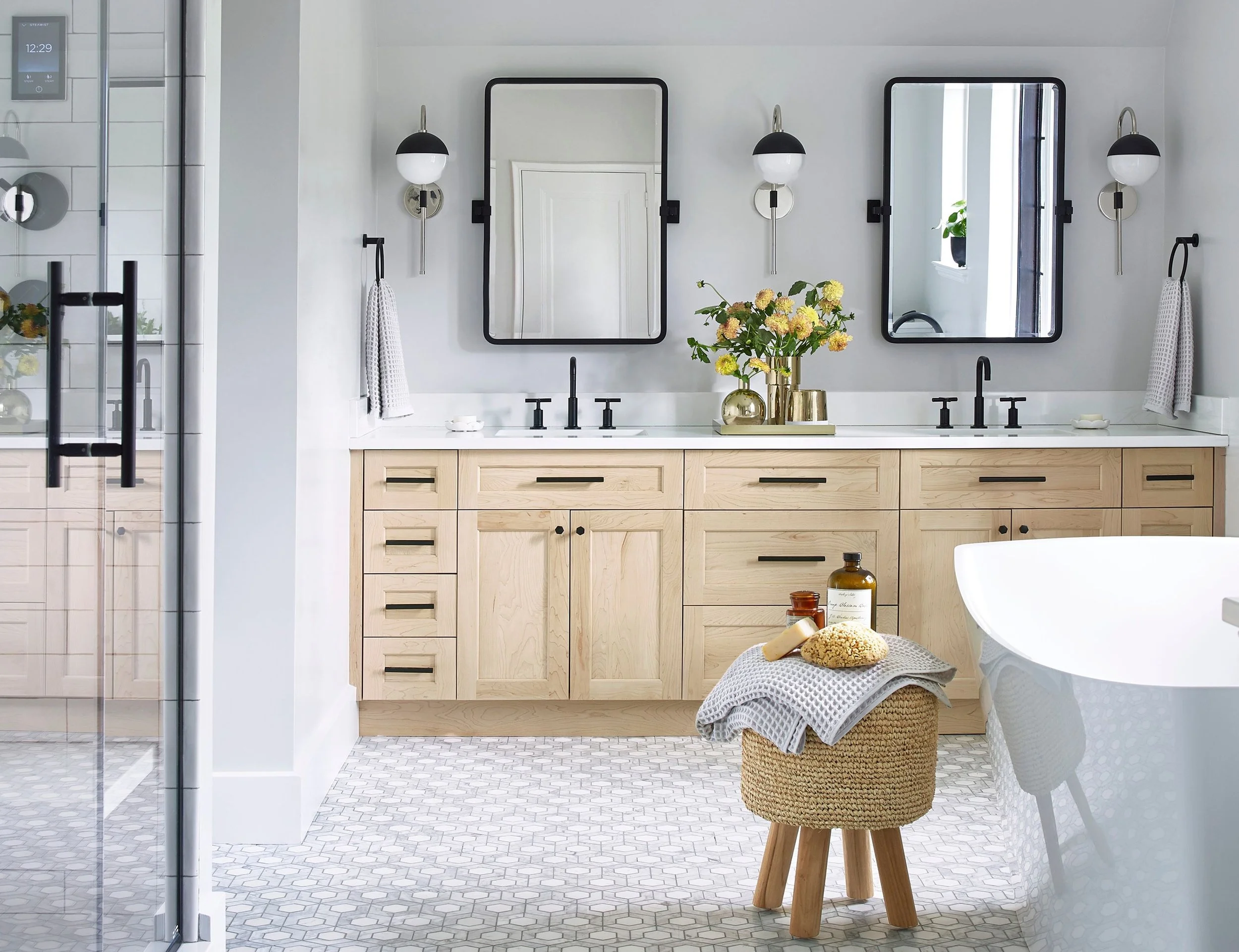 A modern bathroom with a double vanity, two mirrors with black frames, black fixtures, wall-mounted black and white lamps, and a decorative tray with flowers on the countertop. There's a woven basket with towels and bath products in the foreground, and a glass shower enclosure on the left.