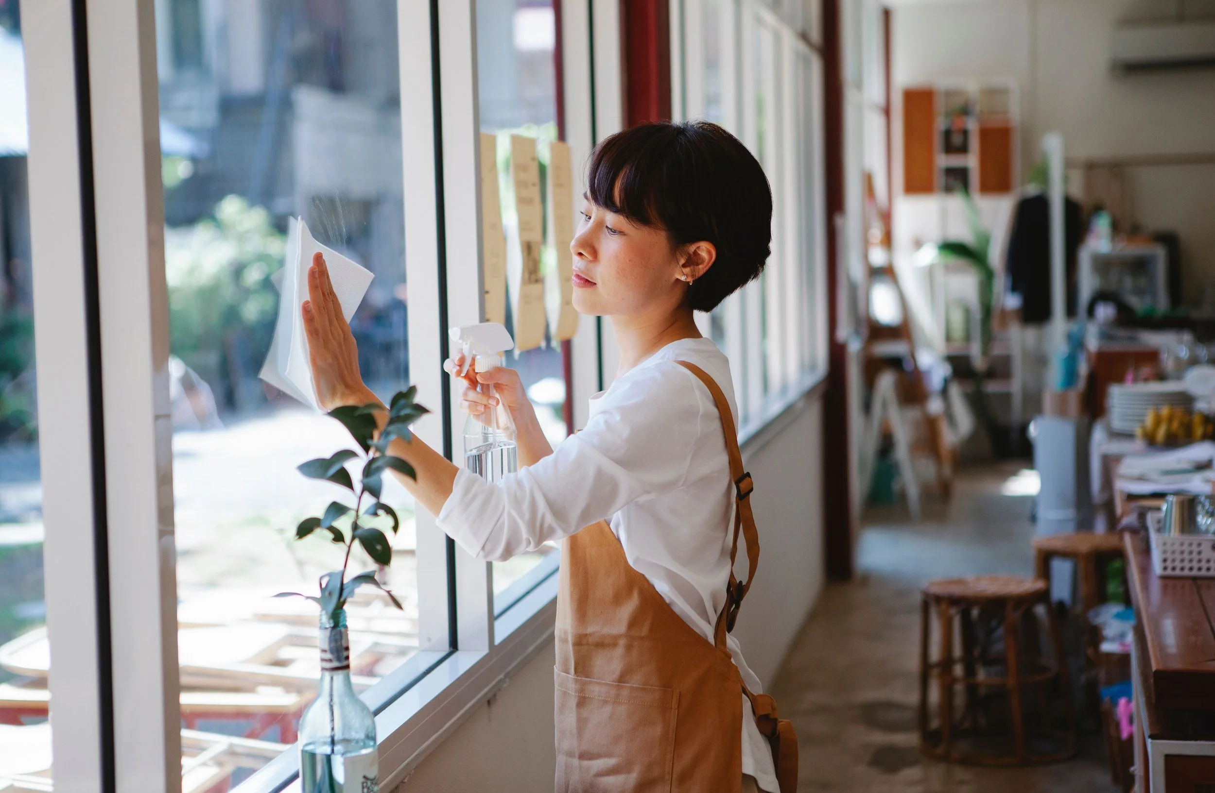 Woman cleaning a large window in a cozy interior space, wearing a brown apron and holding a spray bottle.
