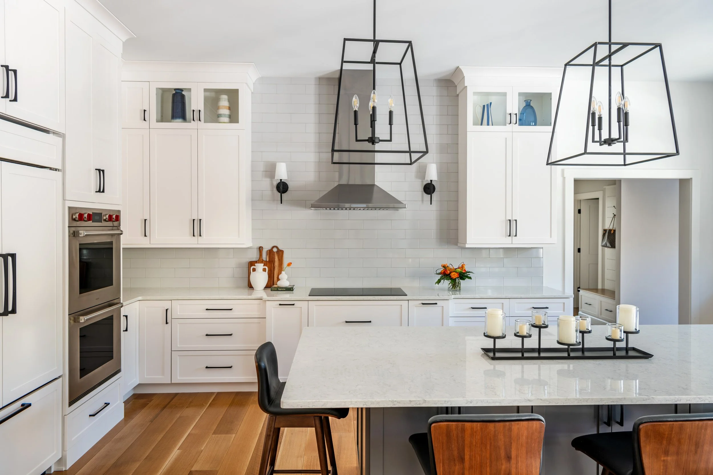 Modern kitchen with white cabinets, stainless steel appliances, black handles, a white countertop, hanging black and glass light fixtures, a flower arrangement, and a black chair in the foreground.