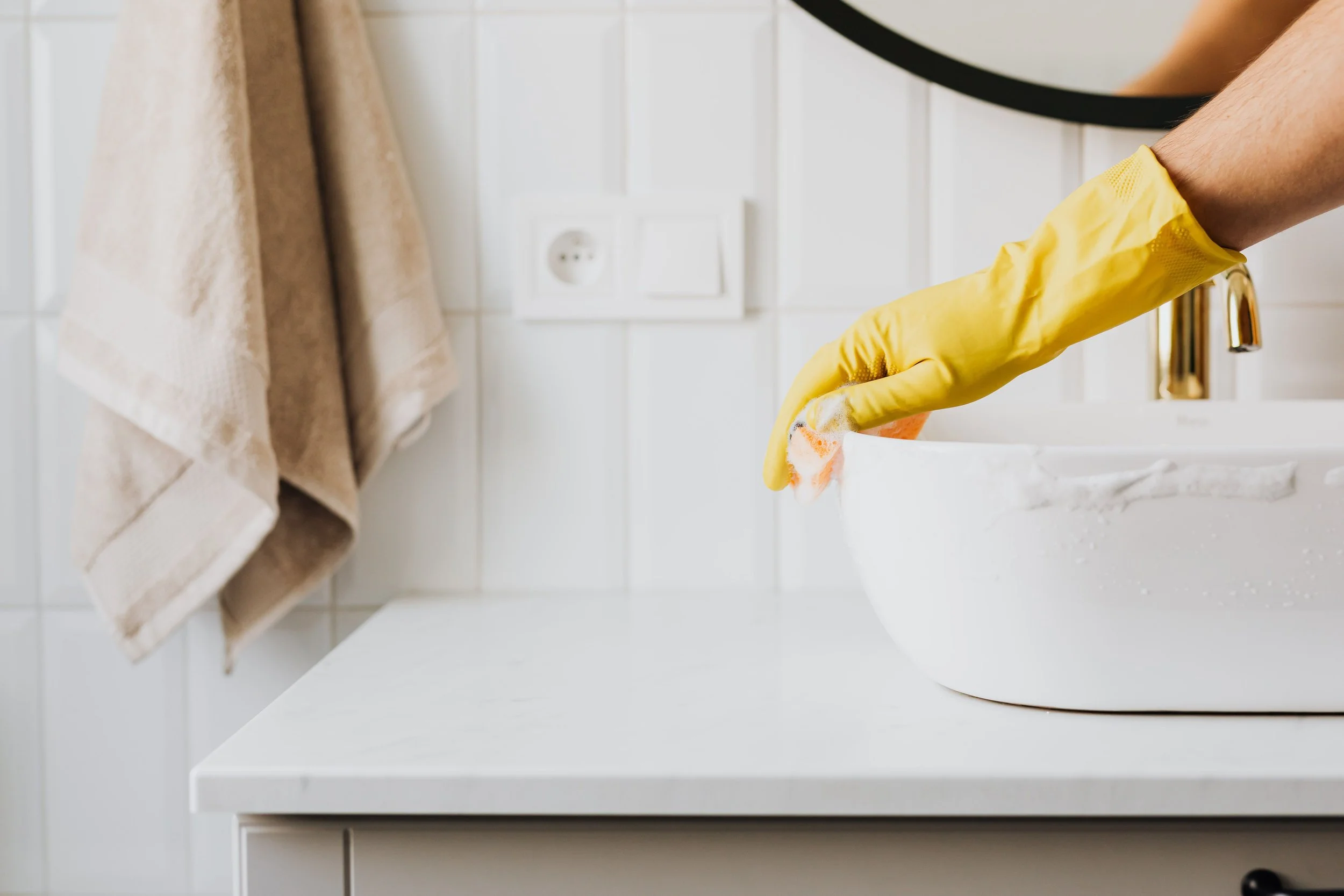 A person wearing yellow rubber gloves cleaning a white bathroom sink with a sponge.
