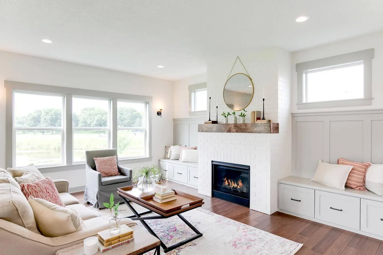 Bright living room with white walls, large windows, a fireplace with a wooden mantel, built-in white benches with cushions, and a beige sofa with pink and beige pillows. A wooden coffee table with books, a glass vase, and a plant. There are recessed ceiling lights and a round mirror above the fireplace.