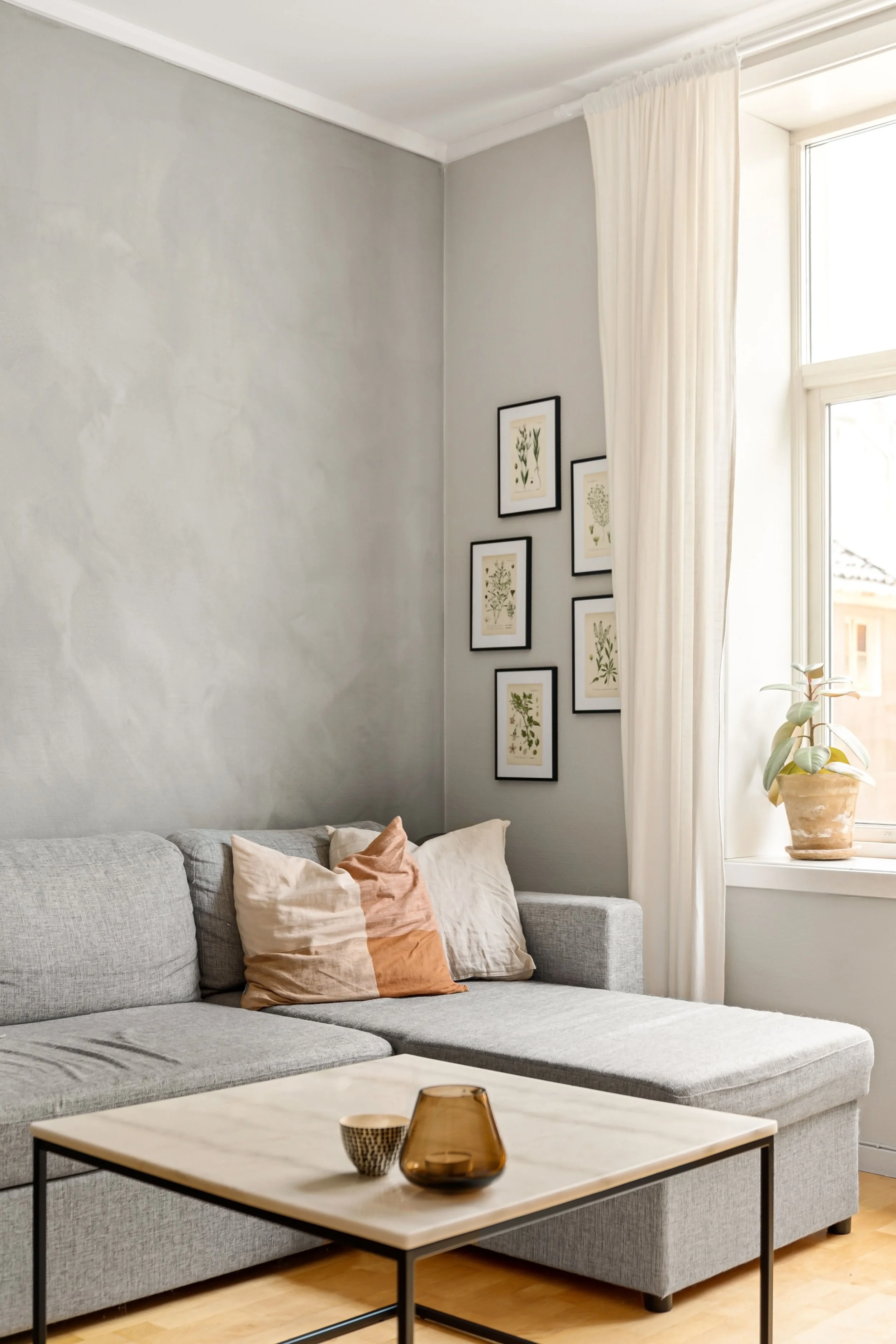 A living room corner with a gray sectional sofa, beige and rust-colored pillows, a coffee table with decorative objects, a wall with framed botanical prints, a window with cream curtains, and a potted plant.