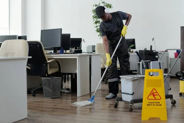 A janitor mopping a wooden office floor in a modern office space with desks, chairs, and computers.