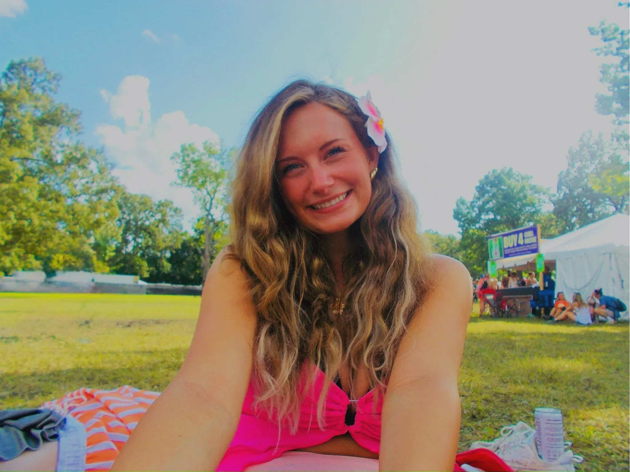 A woman with long wavy hair and wearing a pink dress, smiling outdoors with a blue sky, trees, and a crowd of people at a festival or outdoor event in the background.
