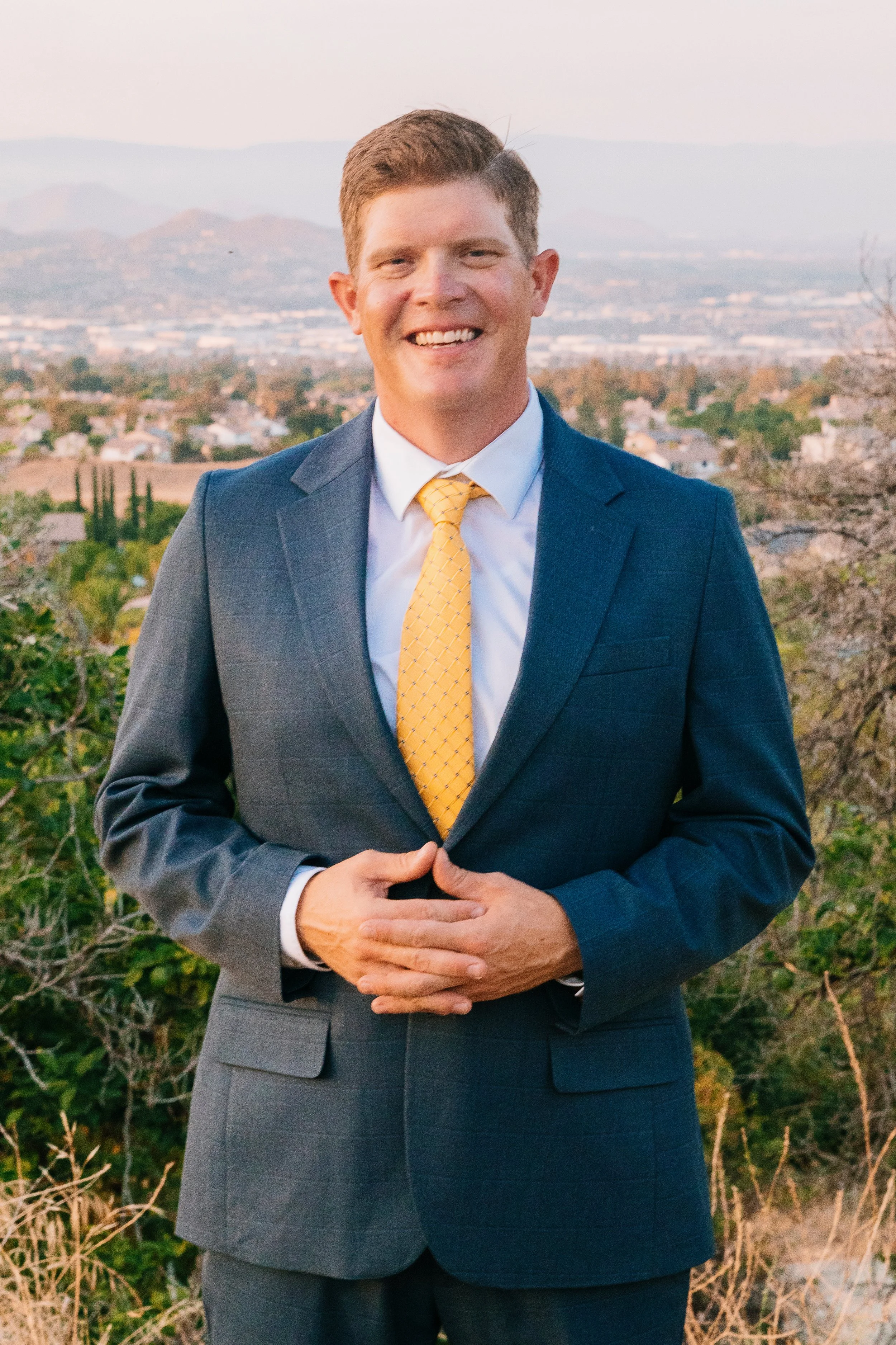 Rory Connell in a dark gray suit and yellow tie smiling outdoors, with a scenic landscape of mountains and Corona, CA, in the background at sunset.