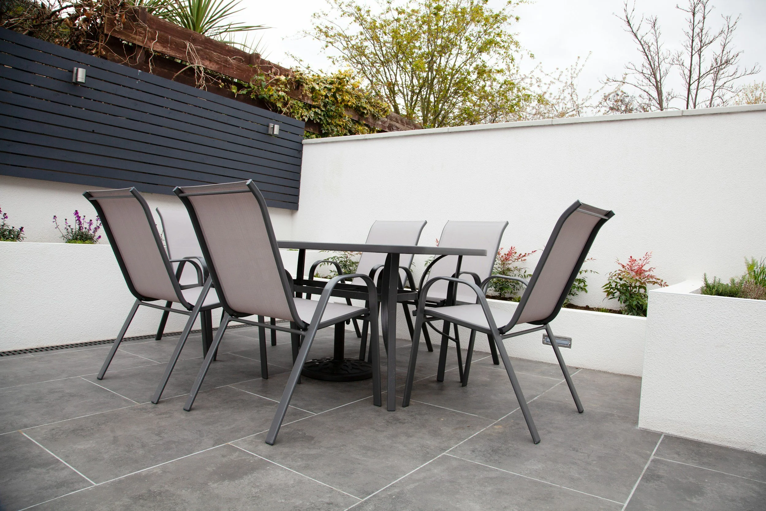 Outdoor patio with black metal table and six gray chairs, white wall with flower garden, and trees with overcast sky.