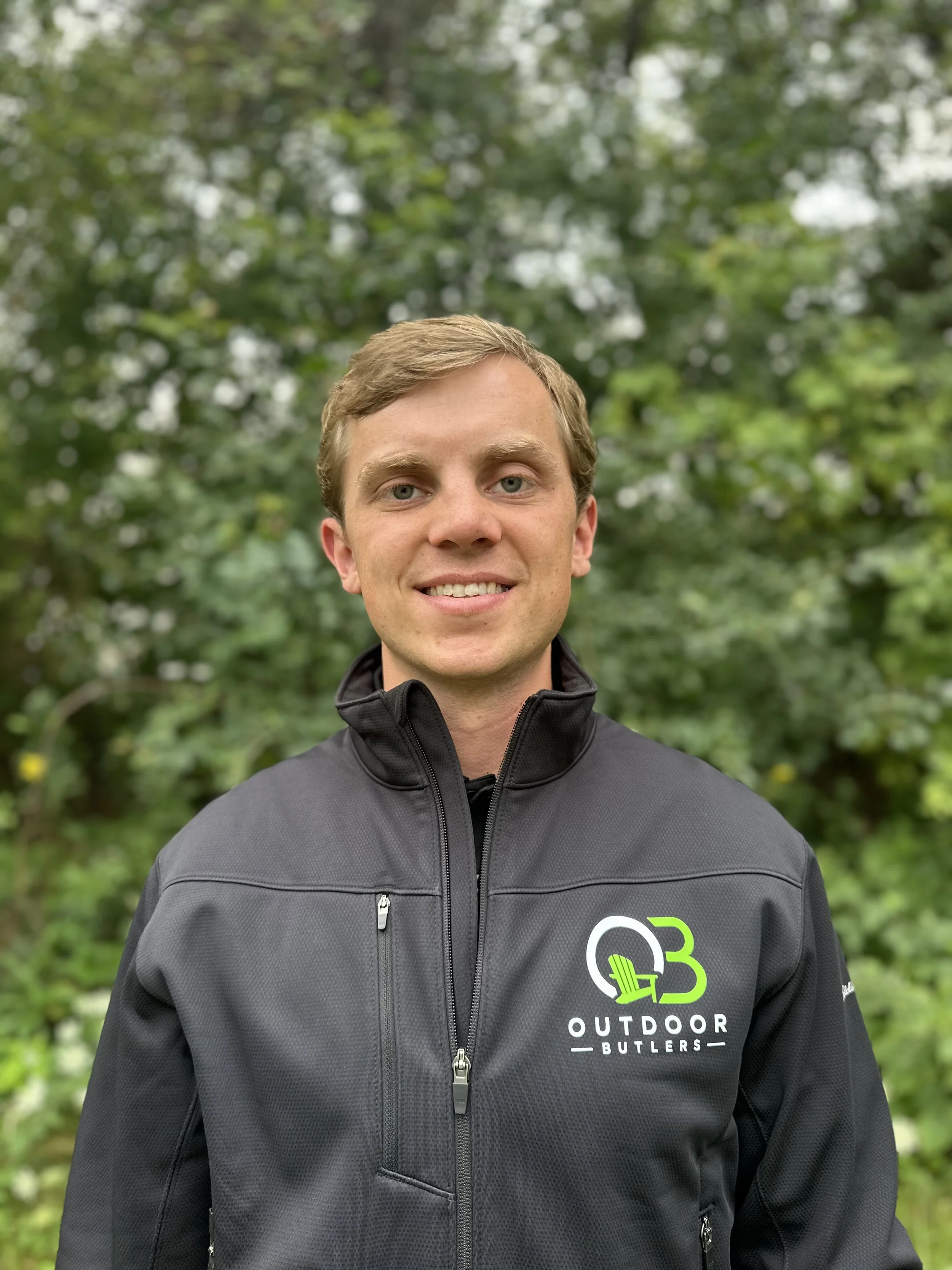 A young man outdoors wearing a black jacket with a logo for 'Outdoor Butlers,' standing in front of a green, leafy background.