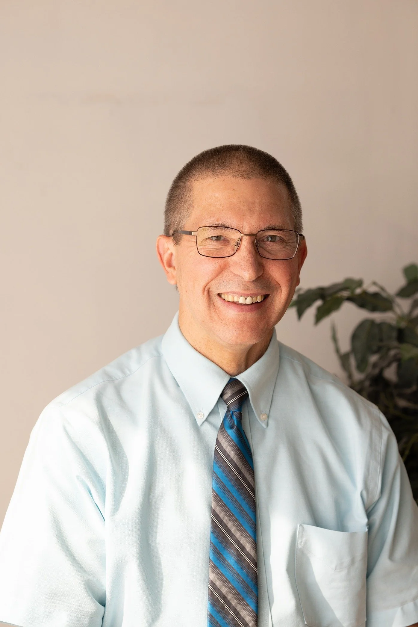Dr. Mike Winkels with glasses, wearing a light blue dress shirt and a striped tie, standing in front of a plain wall and a plant.