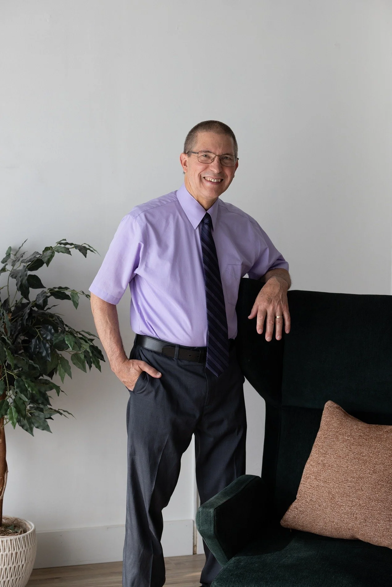 A smiling middle-aged man with glasses wearing a light purple dress shirt, dark purple tie, and dark pants, standing indoors next to a black chair with a brown cushion. There is a potted plant on the left side of the image.