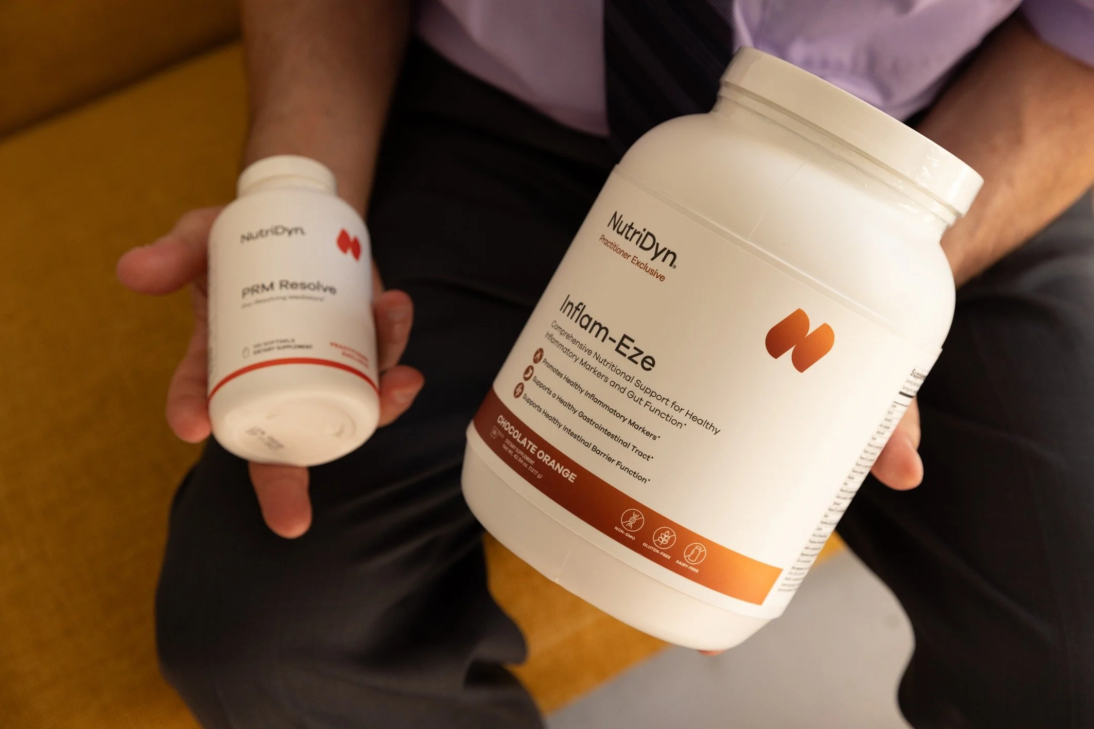 A person holding a white bottle and a white container of dietary supplements. The bottles are labeled NutriDyn, with the smaller bottle labeled PRM Resolve and the larger container labeled Inflamm-Eze, Chocolate Orange flavor.