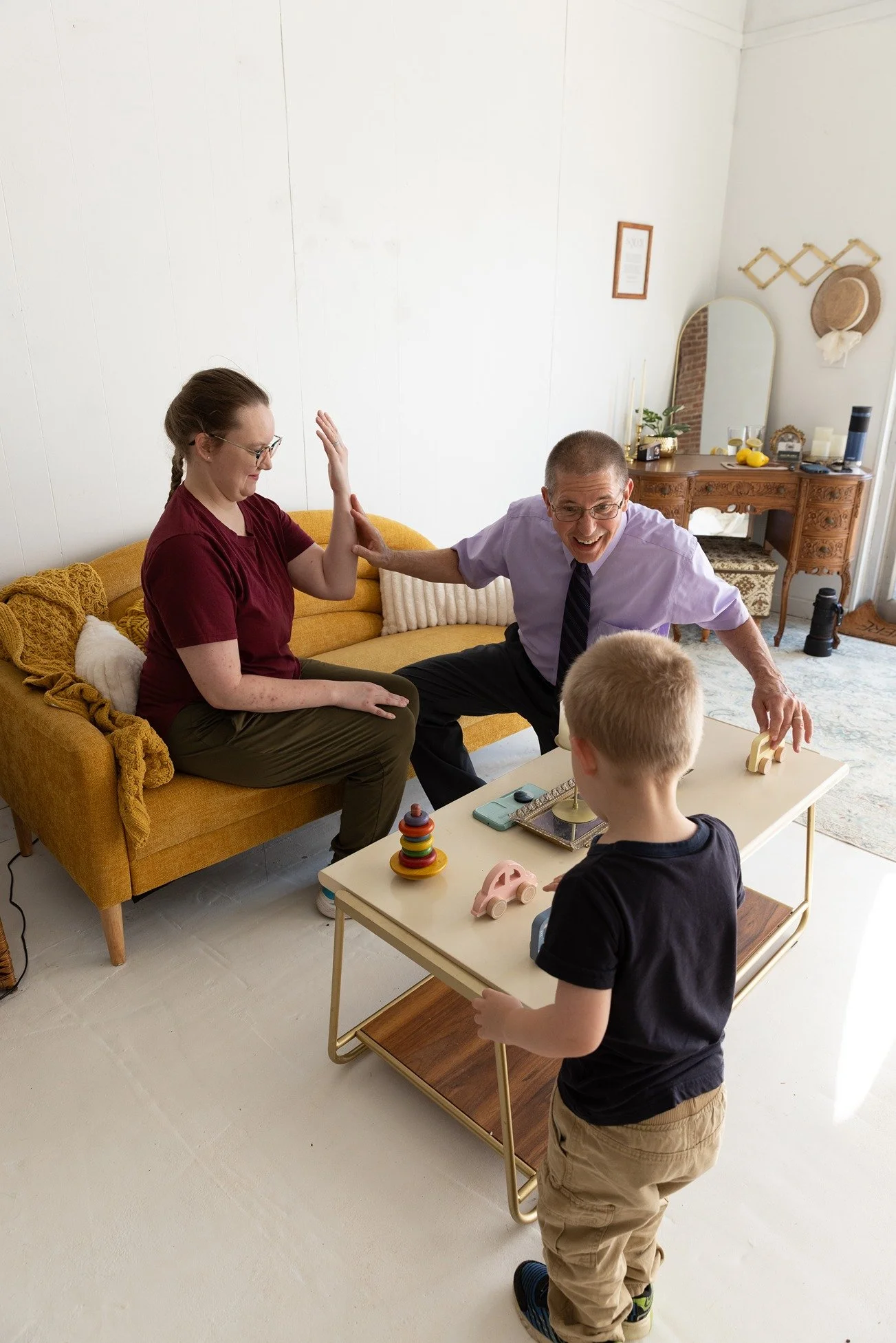 A woman sitting on a yellow sofa giving a high five to a man kneeling next to her. A boy is standing with his back to the camera, facing a toy table with toys on it. The room has white walls and a wooden dresser with a mirror and various decorative items, including candles, a plant, and hats on the wall.