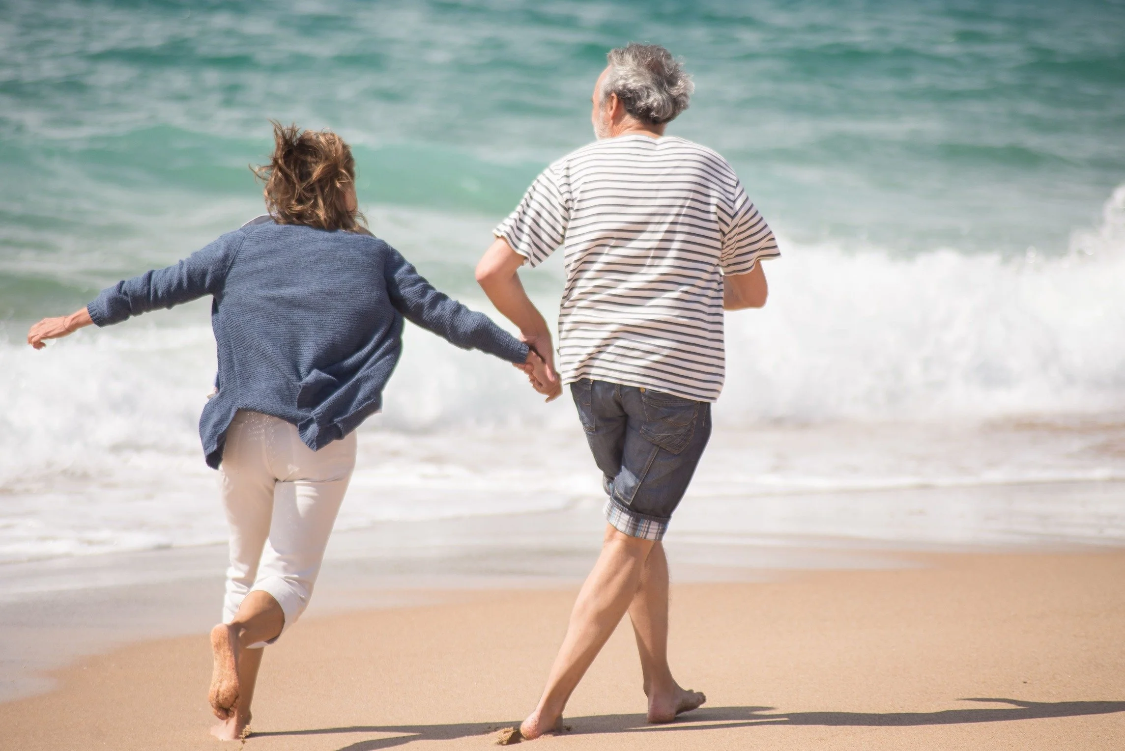 An elderly man and a woman walking on the beach near the shoreline, holding hands, with ocean waves in the background.