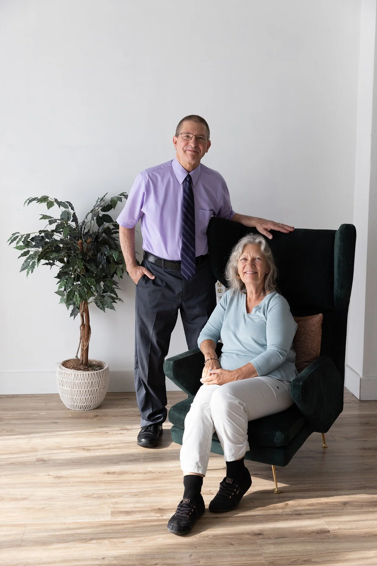 An older woman sitting in a green armchair with blonde curly hair, white pants, black sneakers, and a light blue shirt, smiling. A middle-aged man in a purple shirt, dark pants, tie, and glasses standing beside her, with his hand on the chair's backrest, smiling. There is a tall potted plant on the left and plain white wall background.