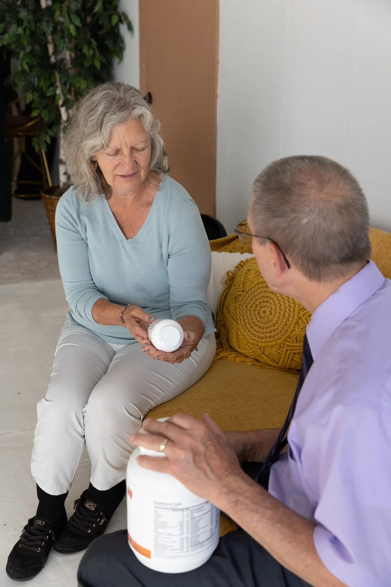 A senior woman with gray hair wearing a light blue top and white pants sitting on a couch, holding a supplement bottle. A man in a light purple shirt and glasses is sitting across from her, also holding a supplement container. They appear to be discussing or examining the supplement bottles.