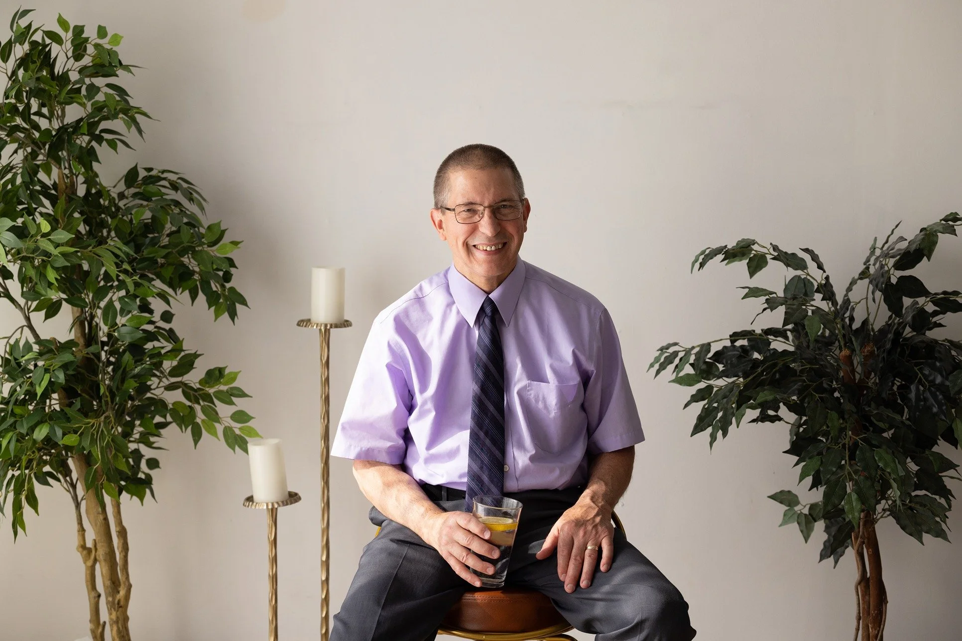 Dr. Mike Winkels with glasses, wearing a purple dress shirt and a dark tie, sitting on a stool, holding a glass of dark-colored beverage, with two large green plants on either side and a white wall background.